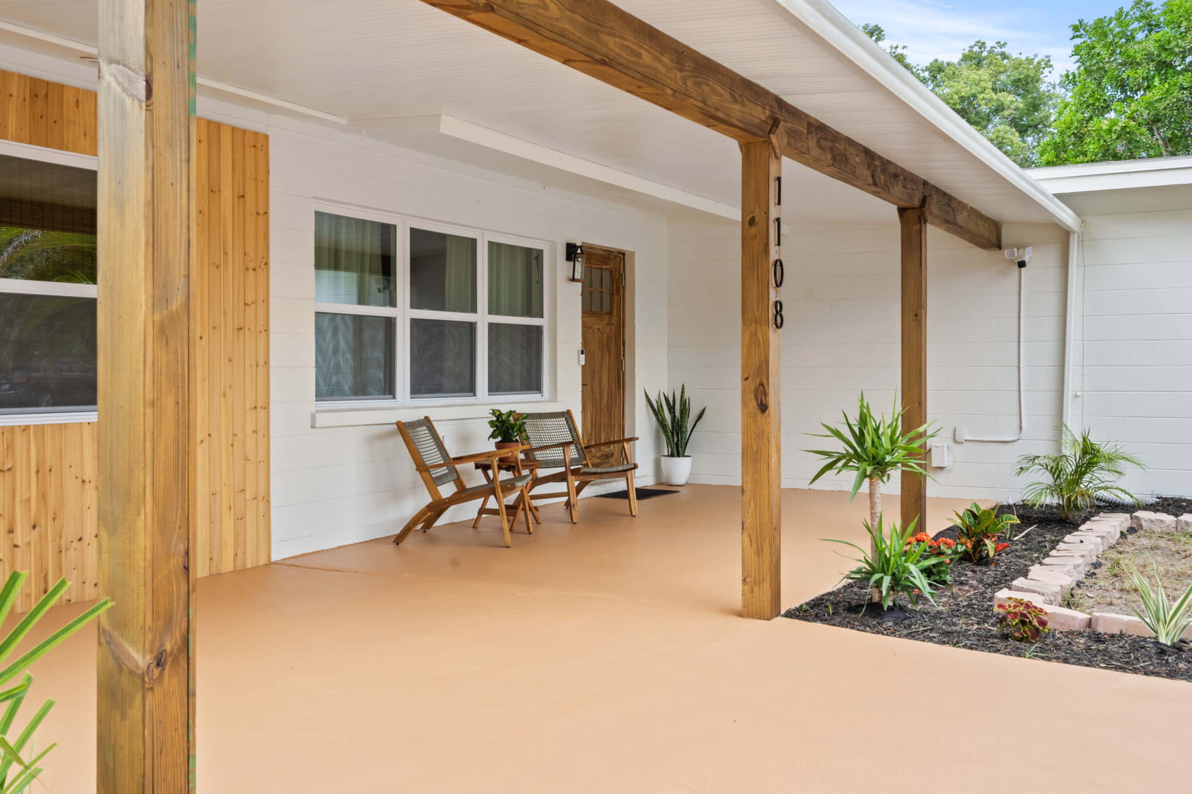 A porch area with a wooden ceiling and support beams, featuring two chairs and a small table on a concrete floor, alongside decorative plants and a well-maintained garden border.