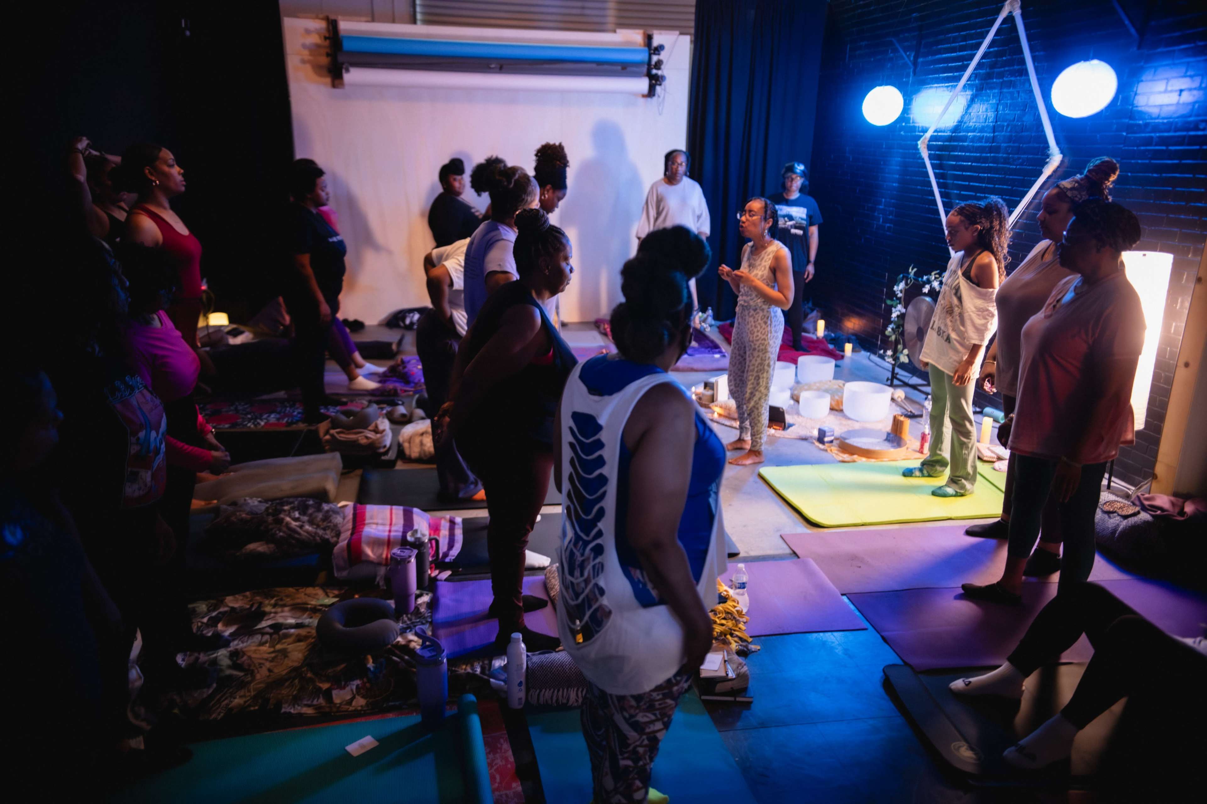 A group of people is gathered in a dimly lit space, participating in a wellness or yoga session with mats and various props scattered around.