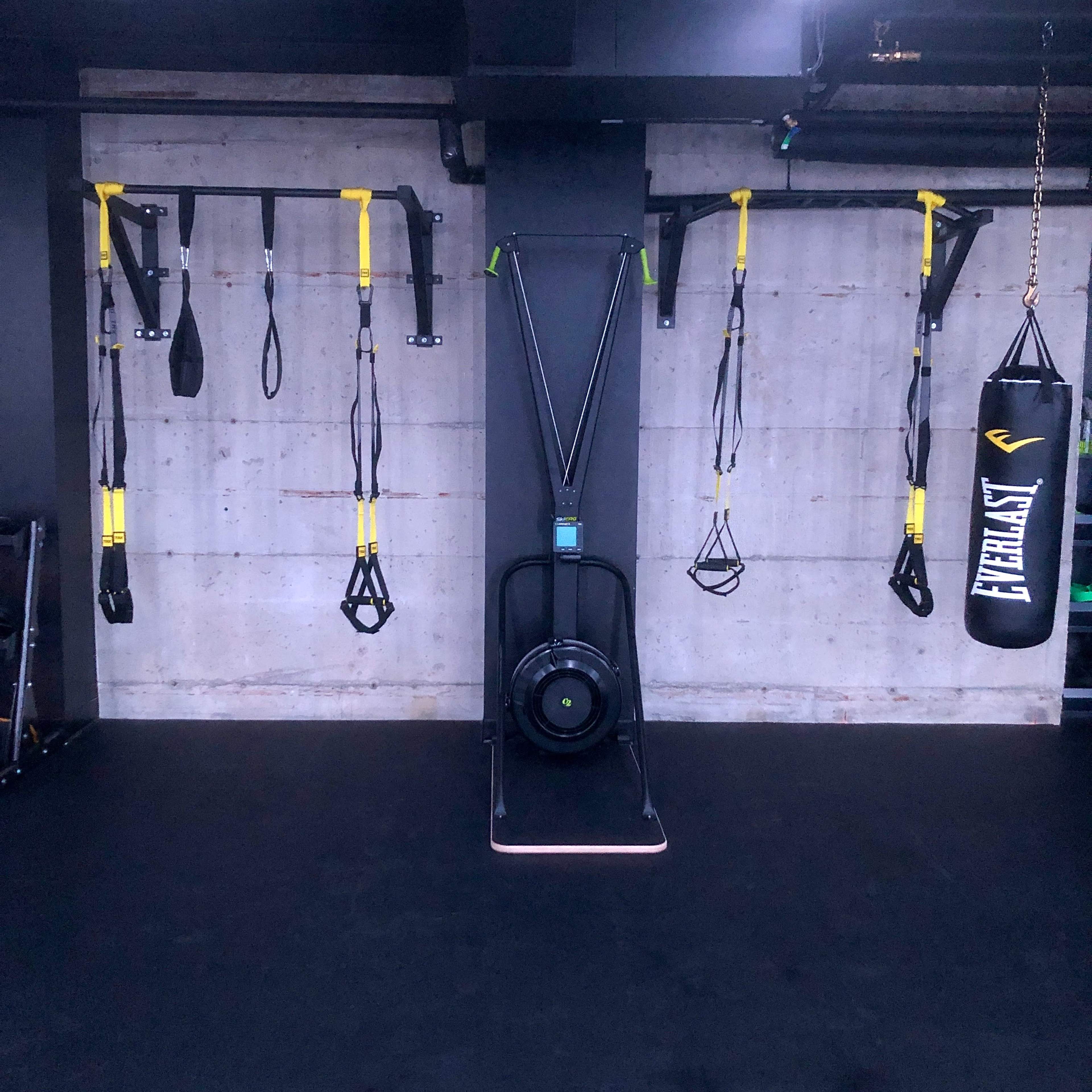 A fitness studio with a rowing machine in the center, surrounded by TRX suspension straps on the wall and a heavy punching bag hanging to the side.