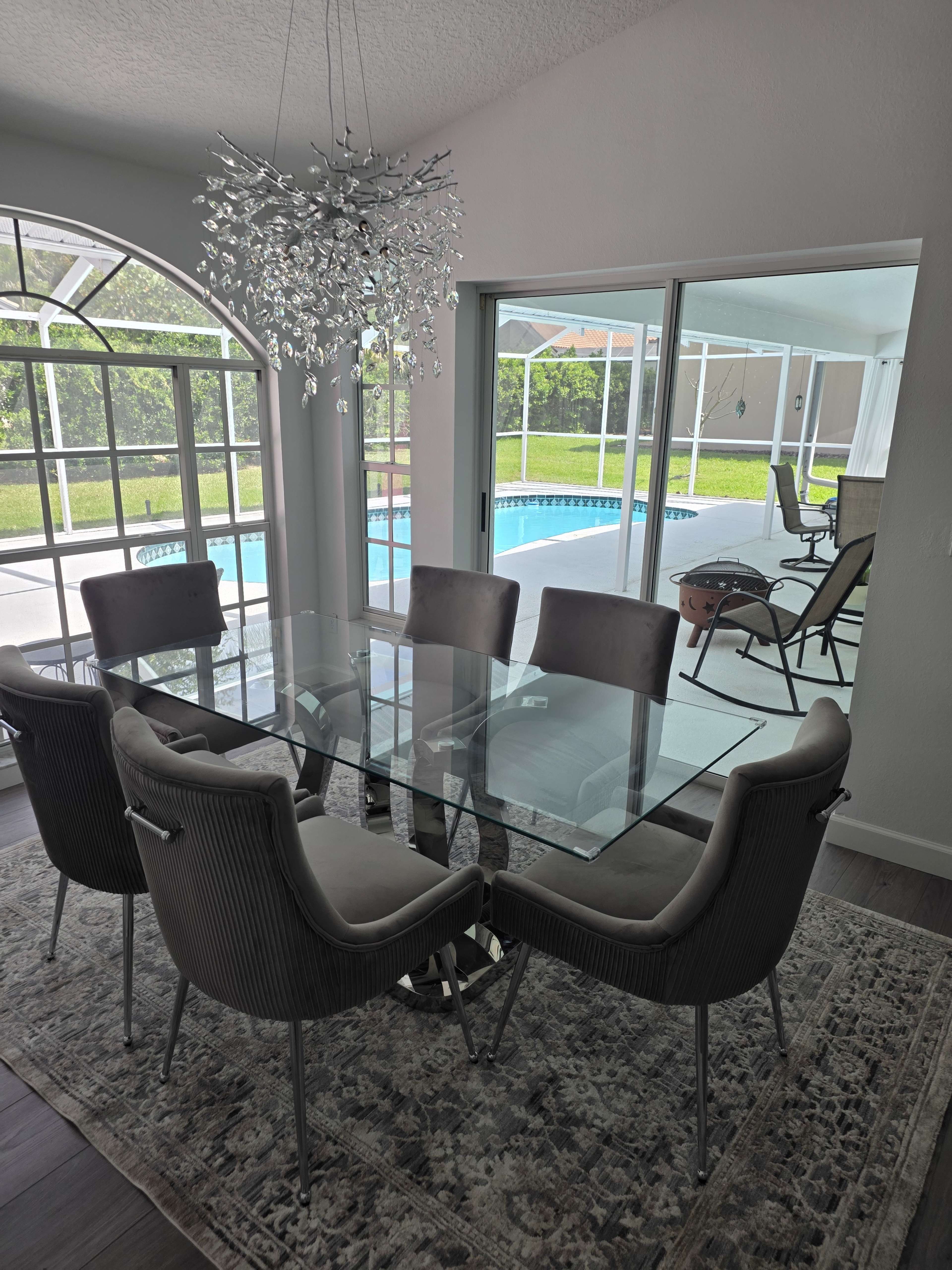 A modern dining area features a glass table surrounded by gray upholstered chairs, with a chandelier overhead and a view of a swimming pool through large windows.
