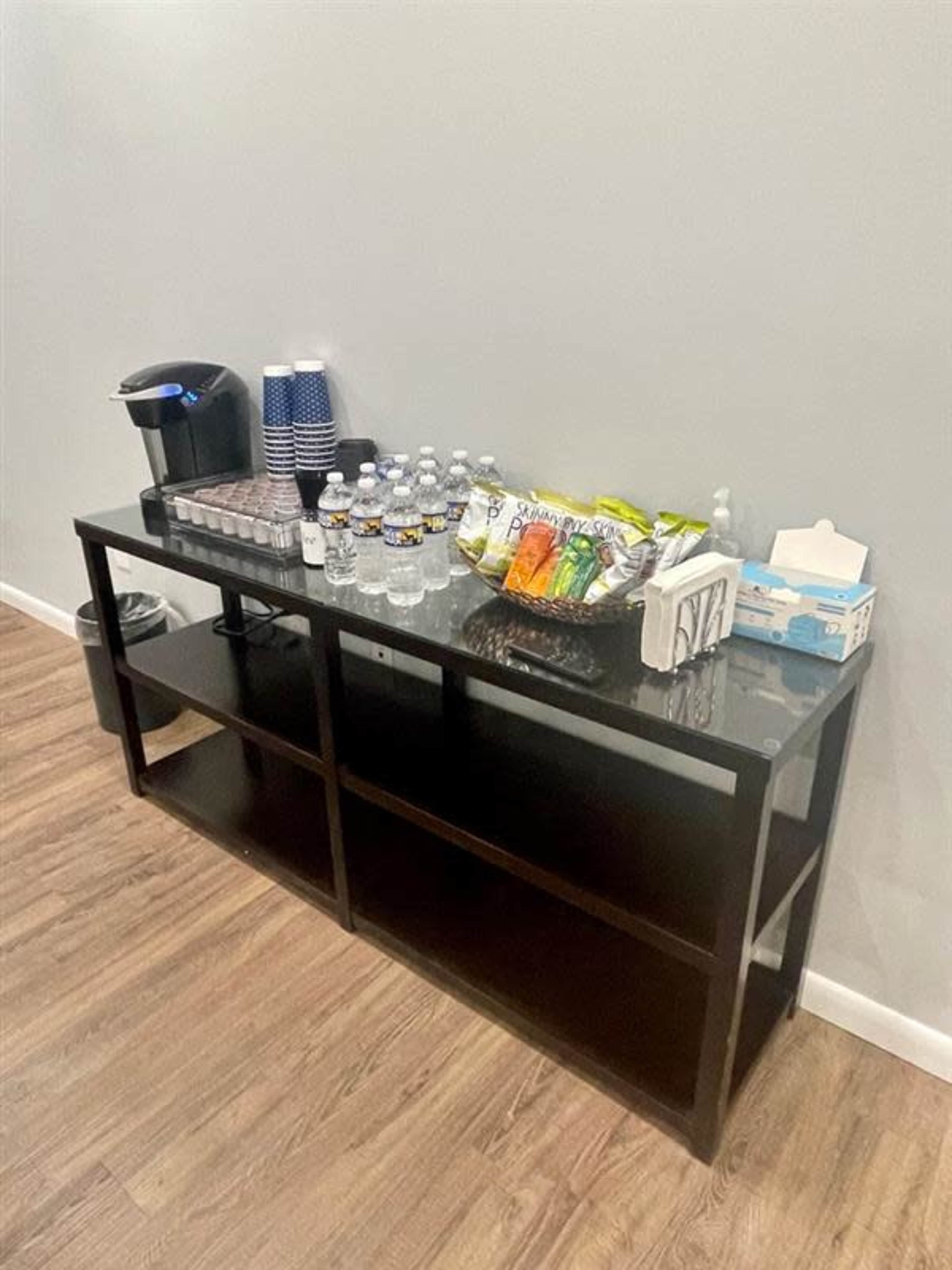 A black wooden table displays a coffee maker, cups, bottles of water, snacks, and tissue boxes against a gray wall.