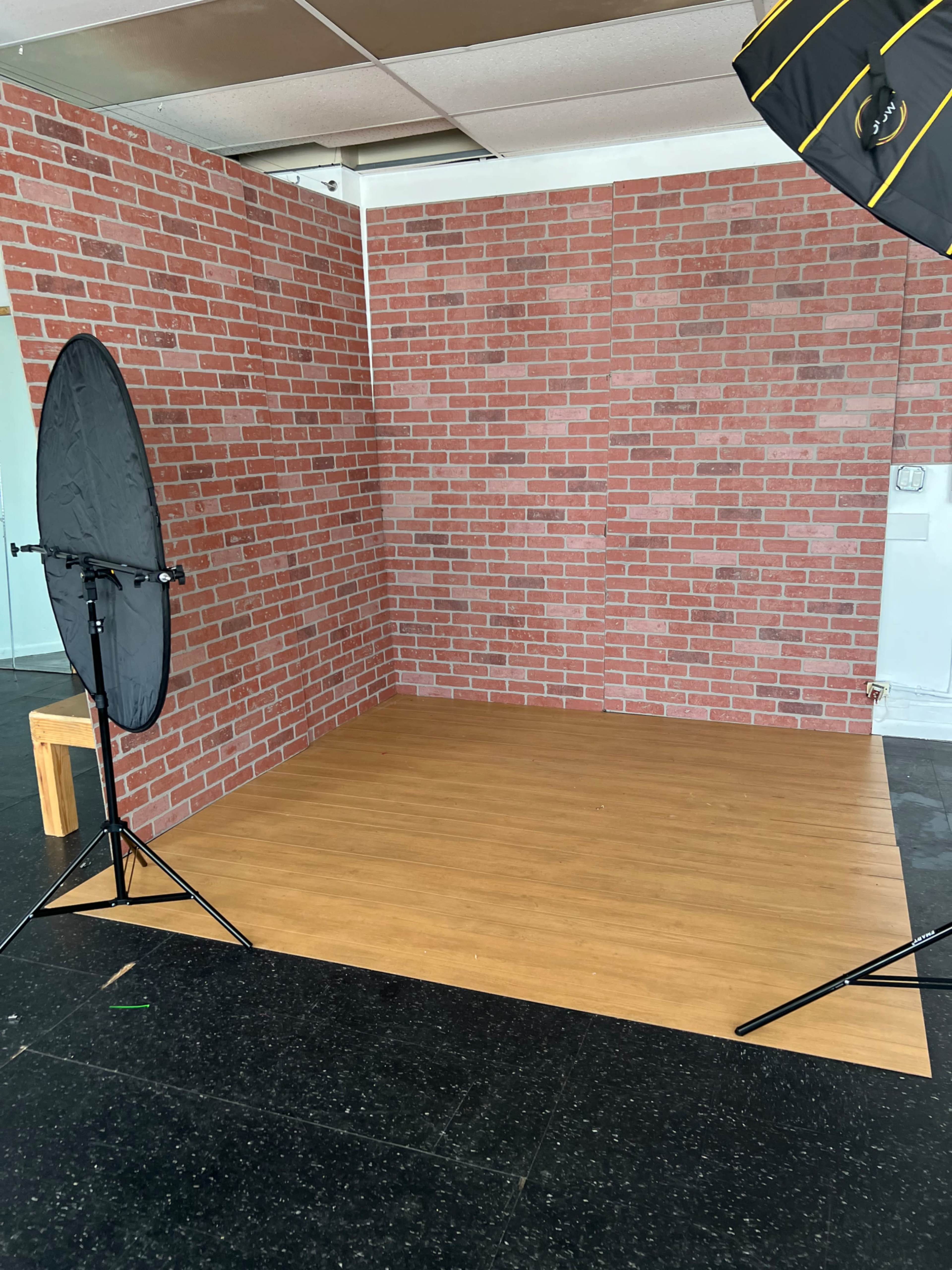 A photography studio with a wooden floor, a faux brick wall backdrop, and two softbox lighting setups.