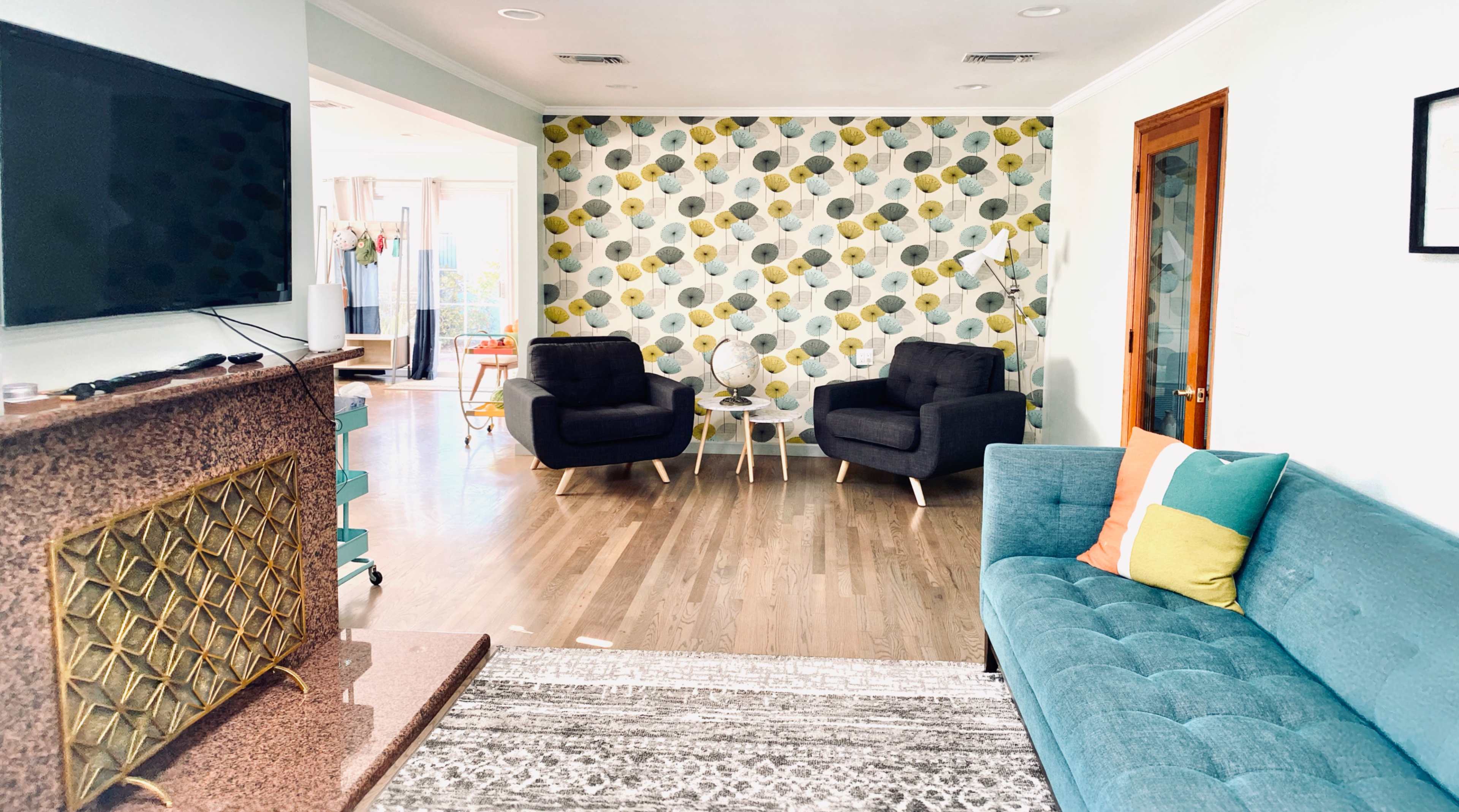 A living room featuring a teal couch, two black armchairs facing a glass table, and a patterned wallpaper with circular designs.