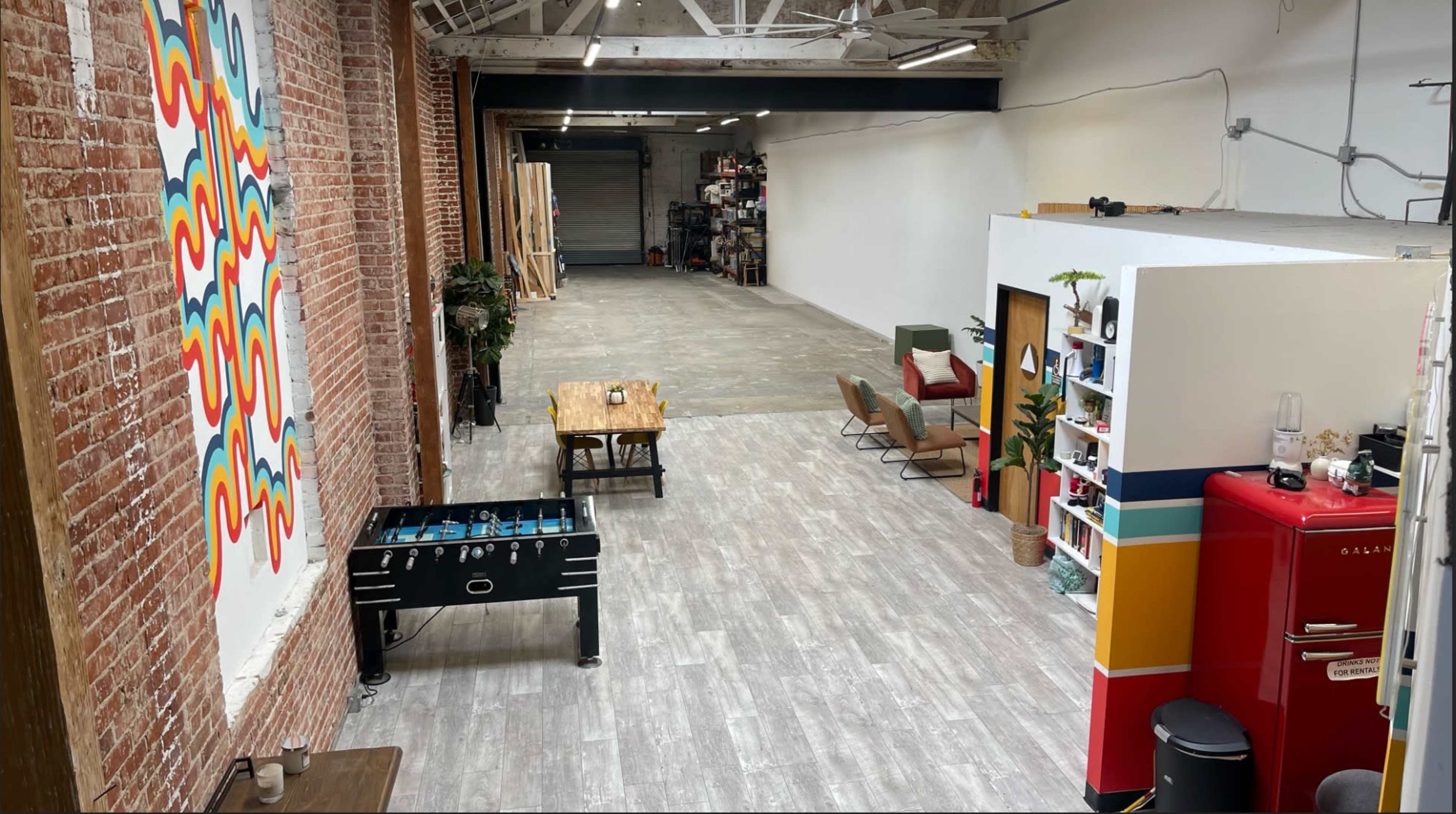 A spacious industrial-style room with exposed brick walls, a foosball table on the left, a dining table in the center, and a bright red refrigerator on the right side.