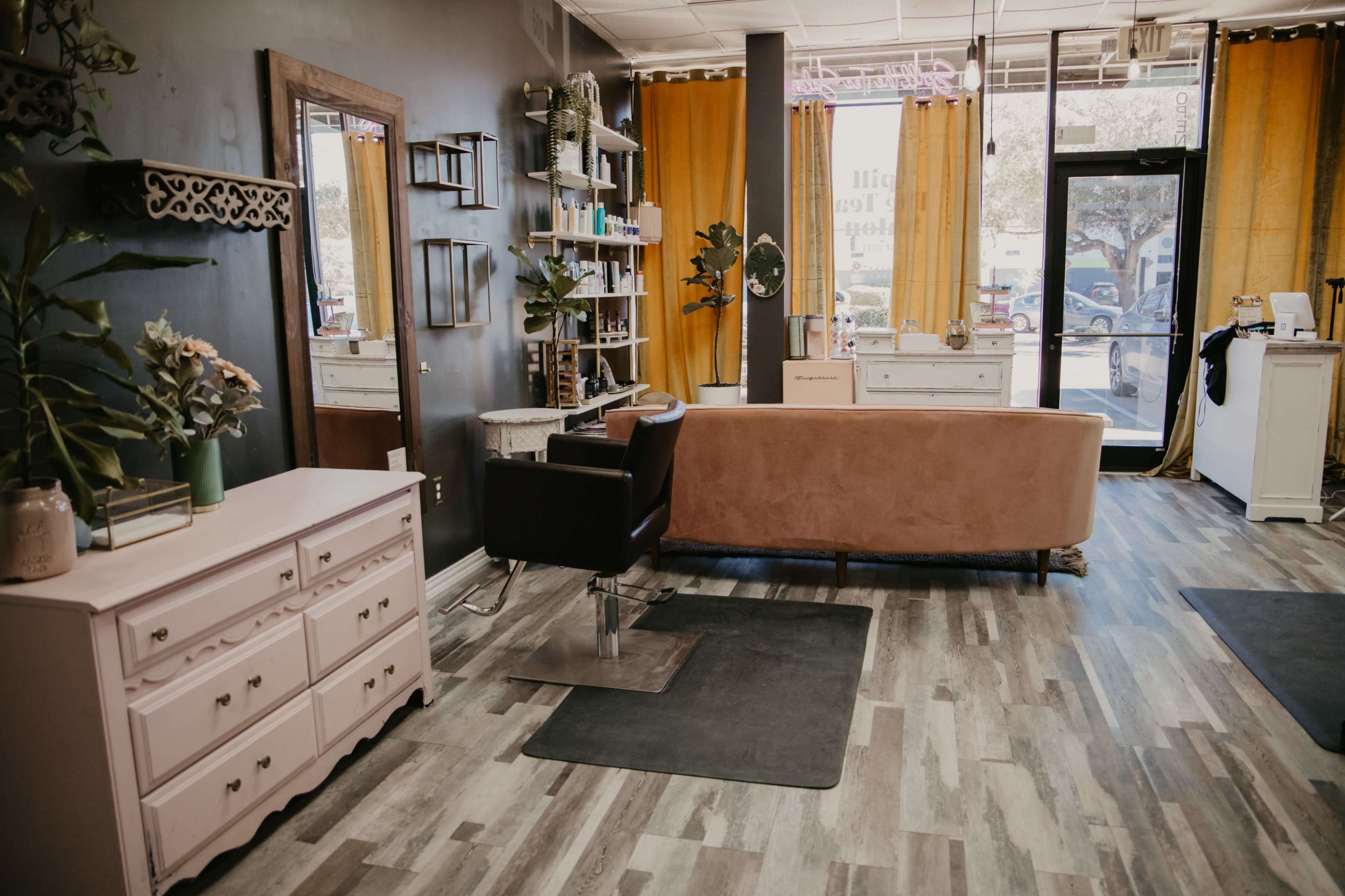 A beauty salon interior featuring a pink upholstered chair, a mirror, shelves with products, and yellow curtains.