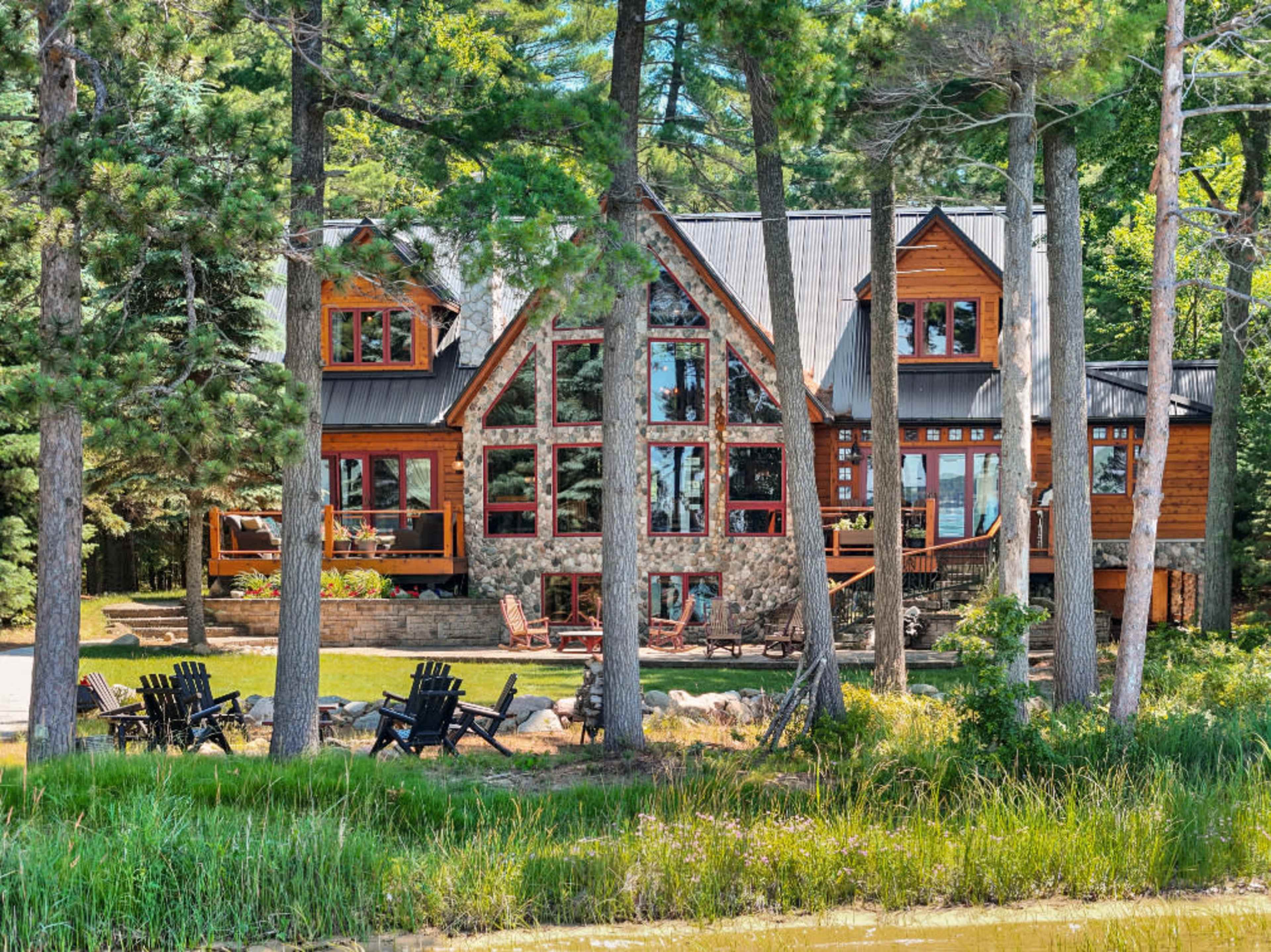 A large, modern cabin made of wood and stone is surrounded by tall trees and overlooks a body of water, with outdoor seating arranged in front.