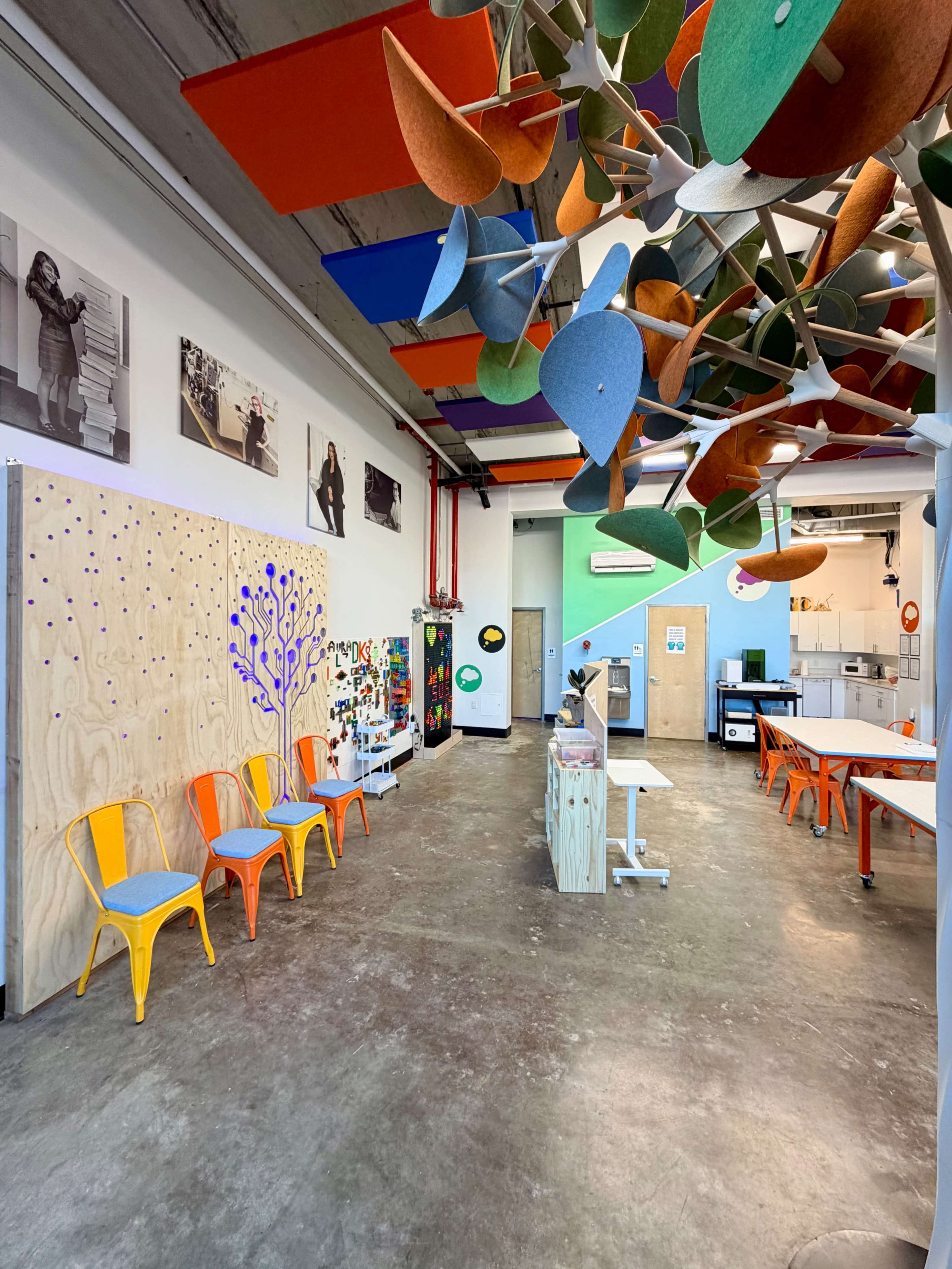 The image shows a brightly colored, modern classroom with wooden paneling, creative decor hanging from the ceiling, and multiple workstations arranged throughout the space.