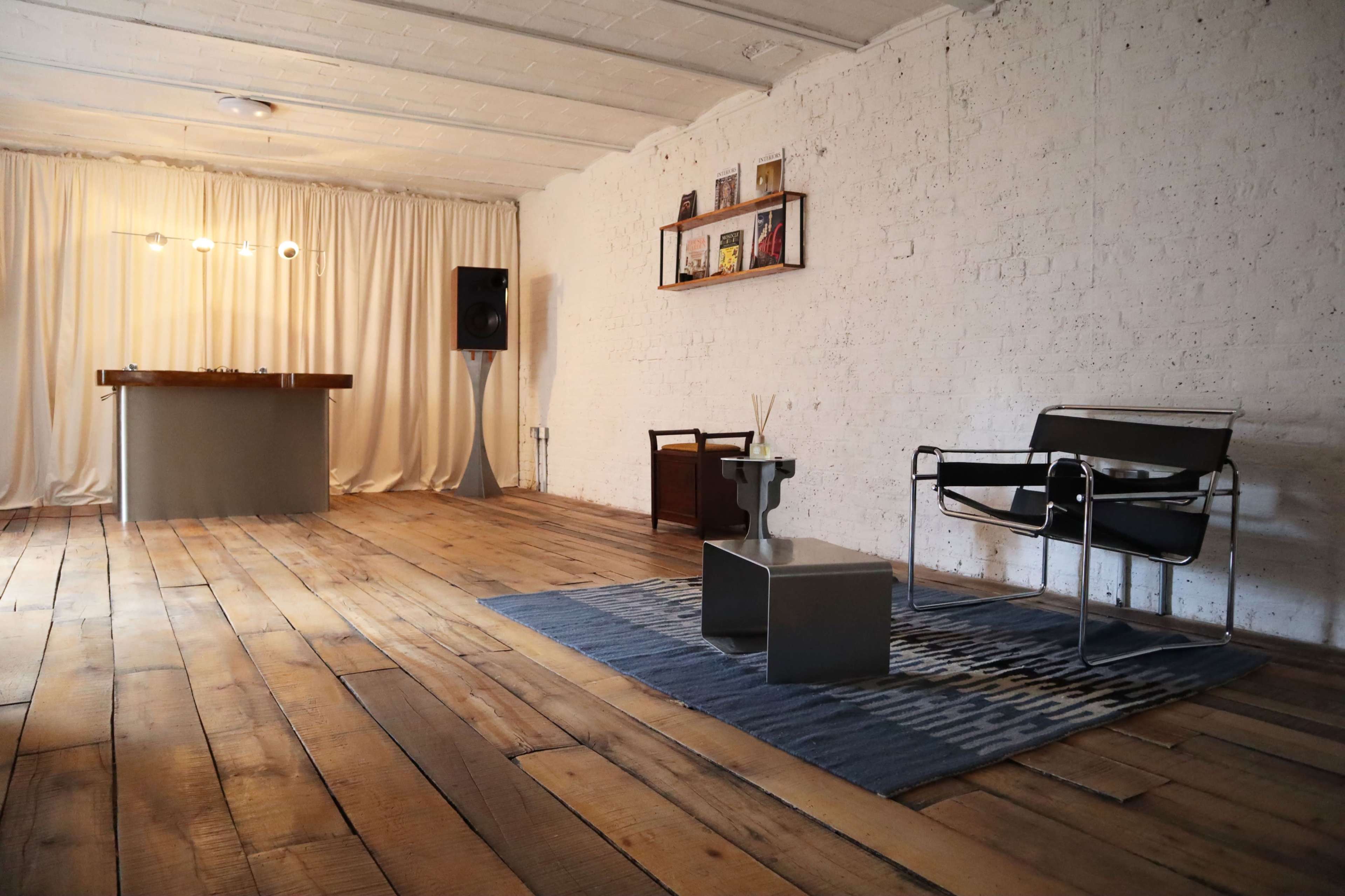 The image shows a spacious, minimalist room with wooden floors, featuring a small table, a chair, and a backdrop of curtains and shelves on one wall.