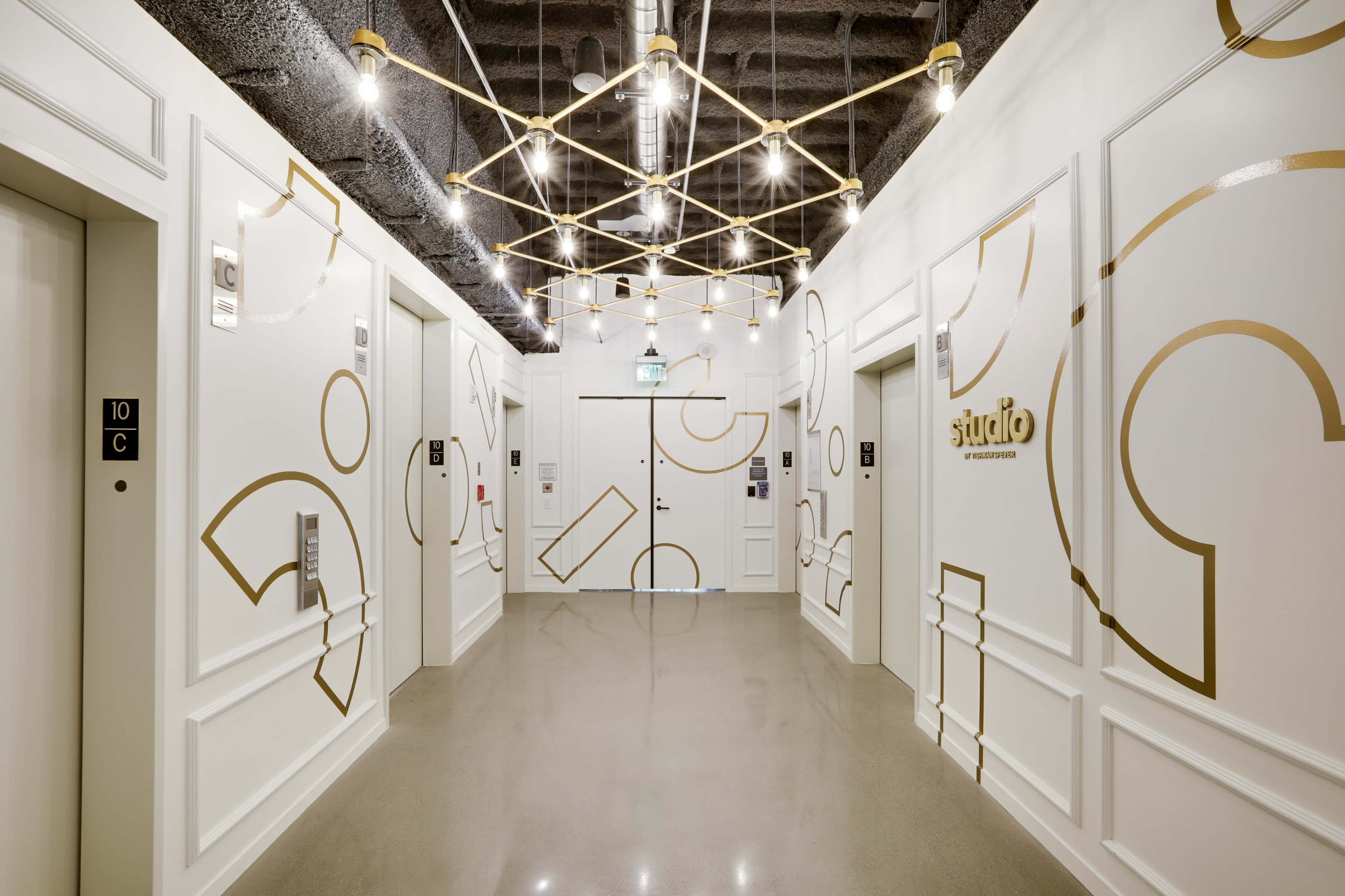 A bright, modern hallway featuring geometric wall designs, with elevators on either side and a decorative ceiling light structure.