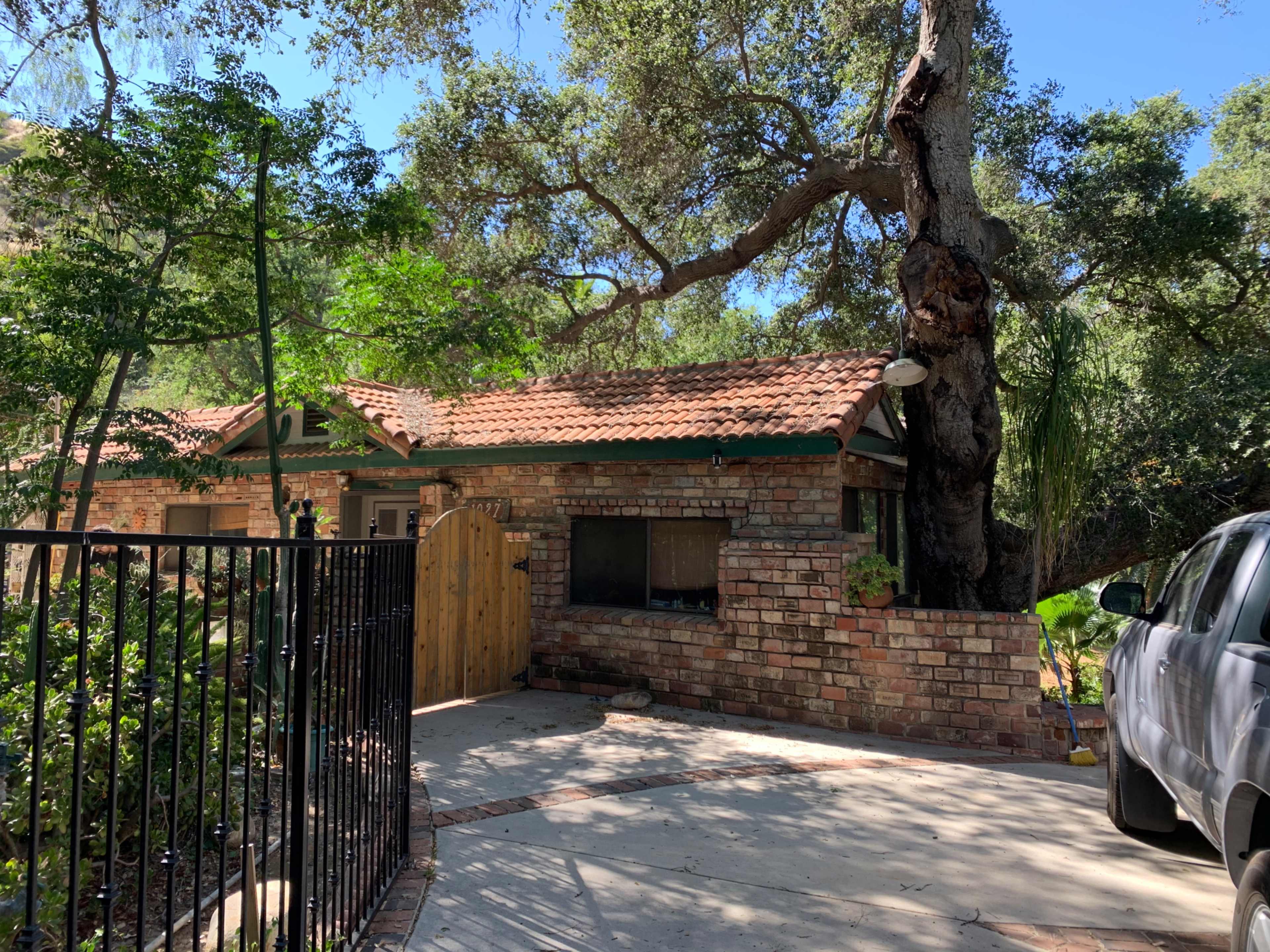 A brick house with a red tile roof sits under large trees, with a gated entrance and a white truck parked in the driveway.