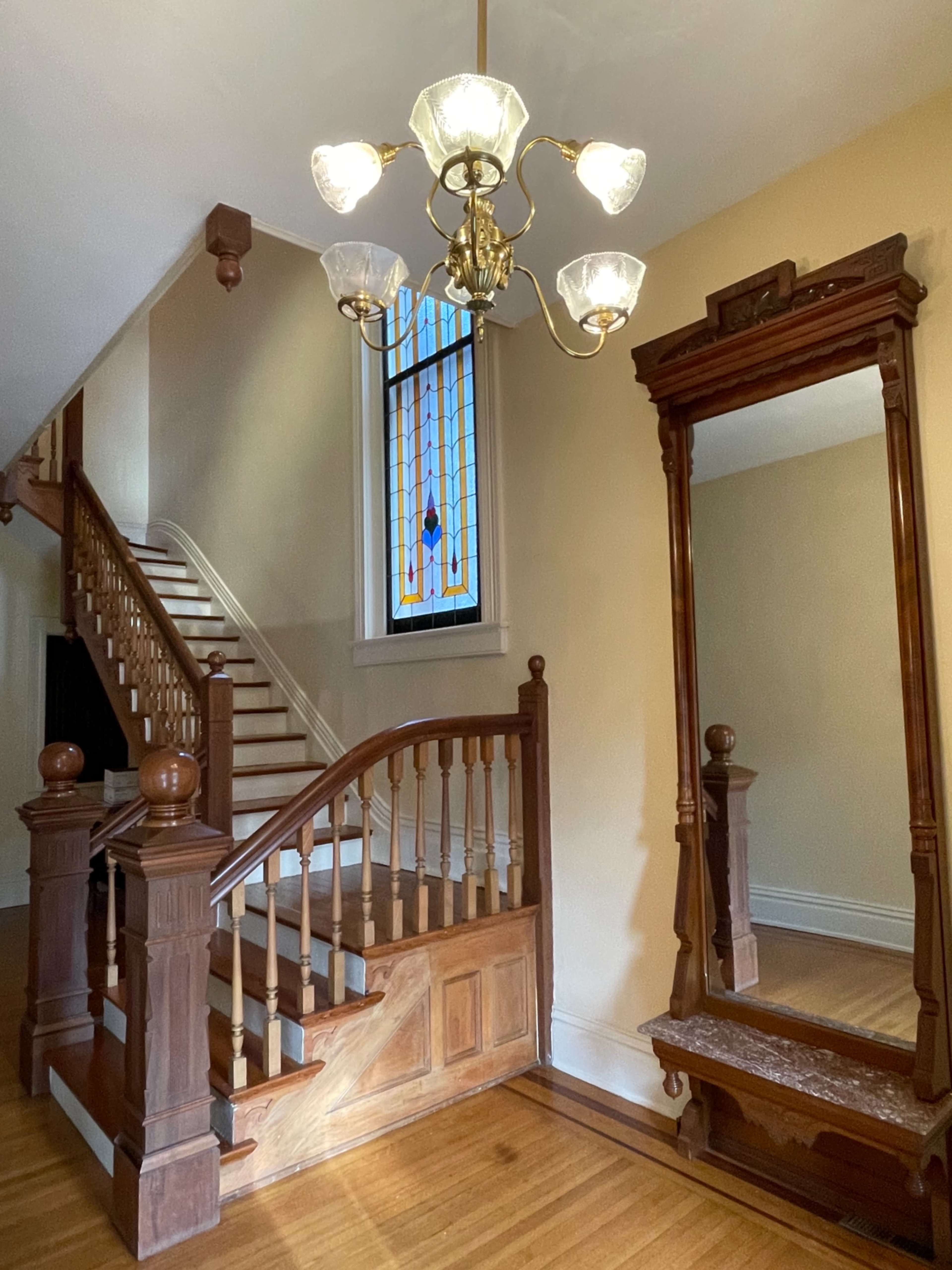 A wooden staircase with a landing, a chandelier, and a decorative mirror, beside a stained glass window.
