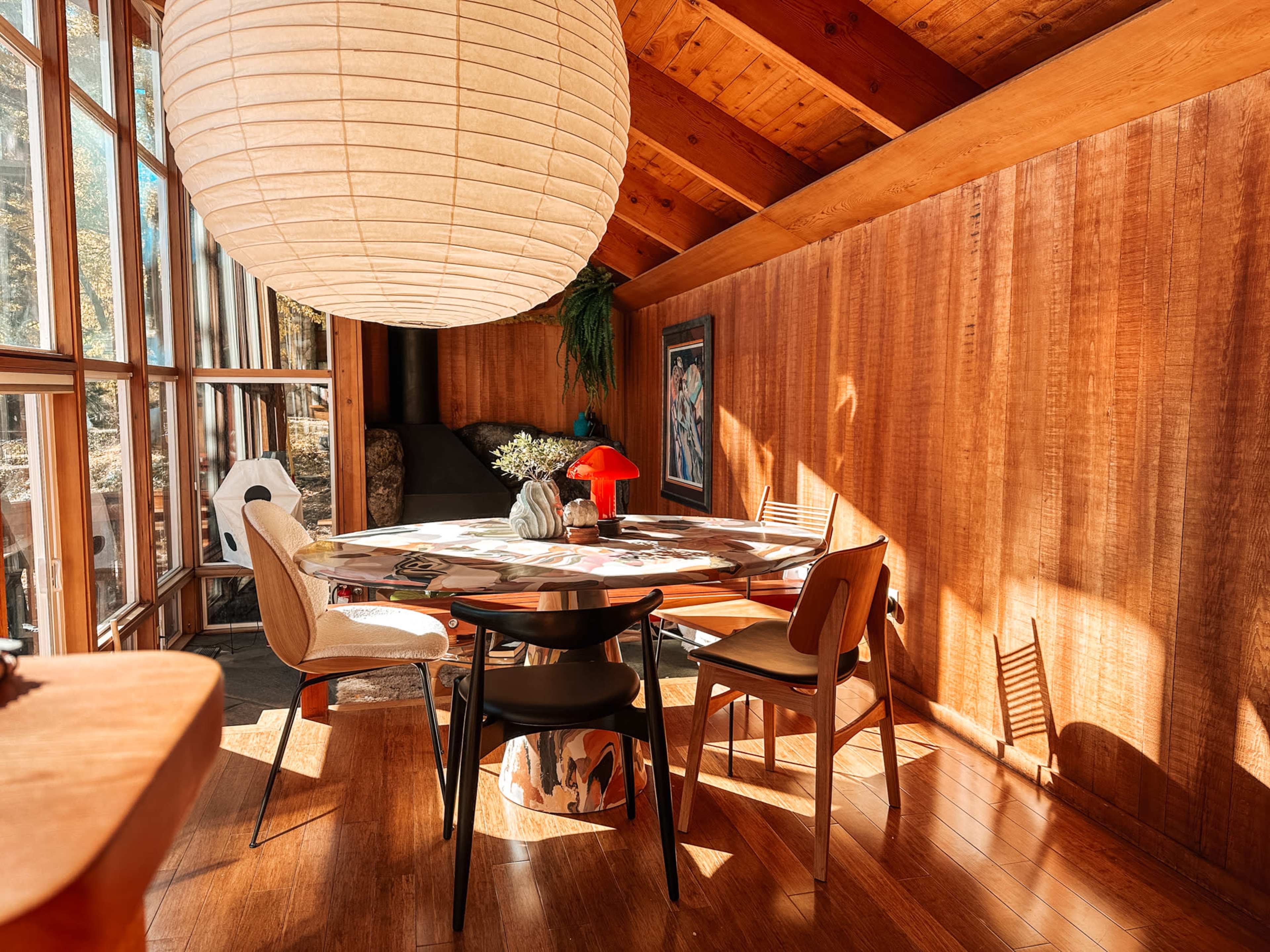 A round dining table with chairs is illuminated by a large paper lantern, set in a wooden room with large windows and a warm ambiance.