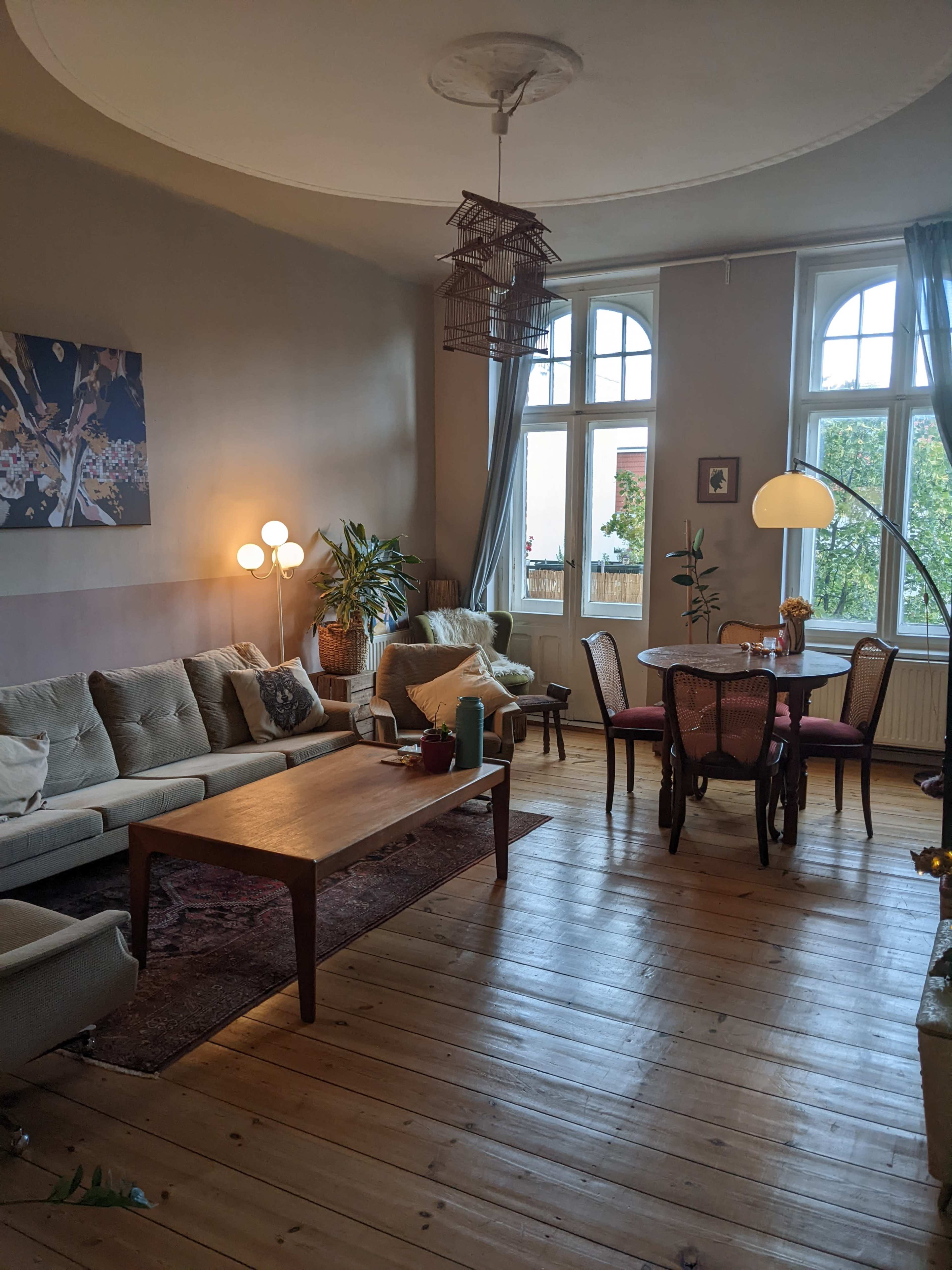A spacious living room features a light-colored sectional sofa, a wooden coffee table, a dining area with wicker chairs, and large windows allowing natural light.