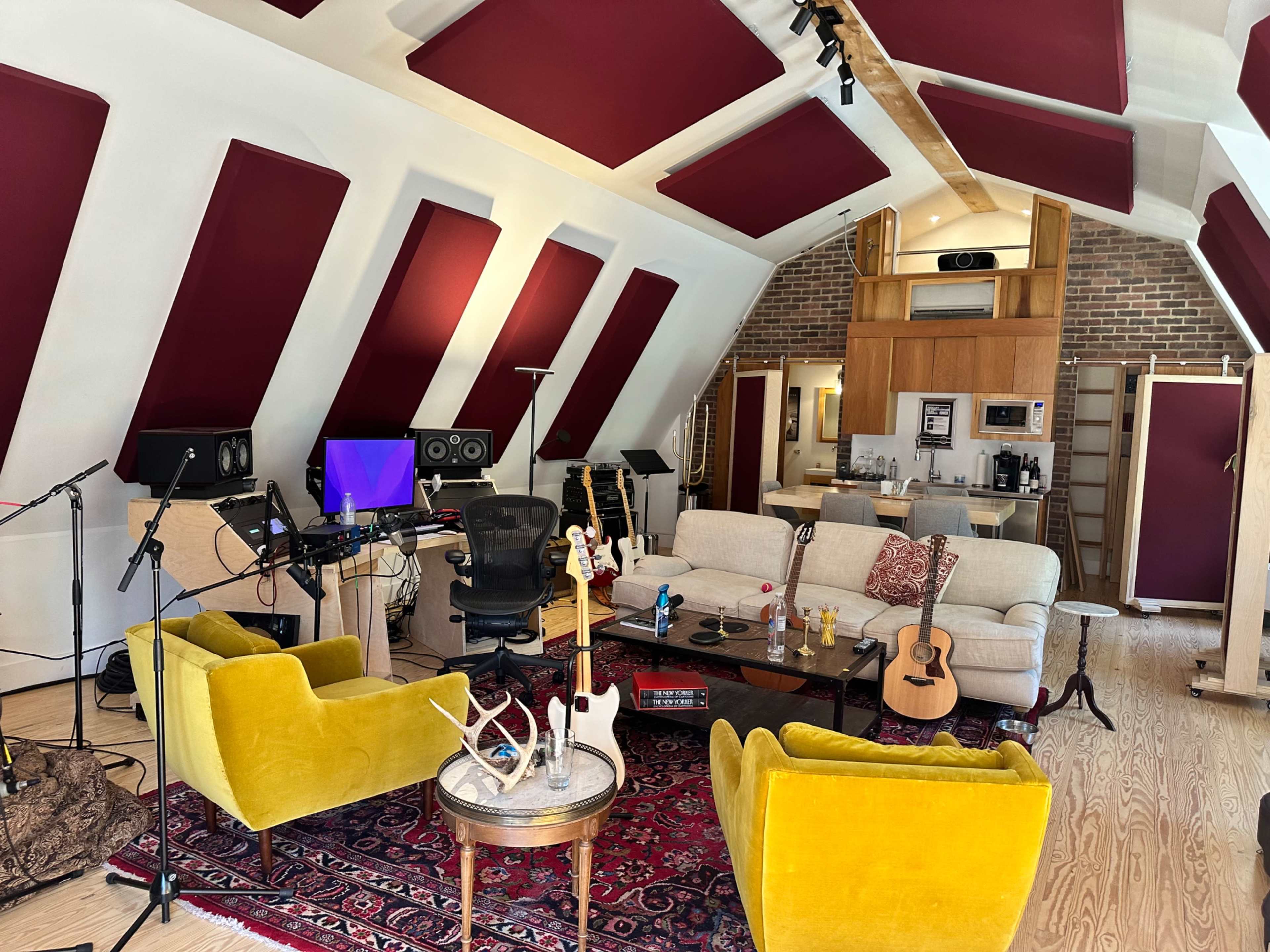A music studio with a central seating area featuring yellow armchairs, a sofa, musical instruments, and sound equipment against a backdrop of angled walls and a kitchenette.
