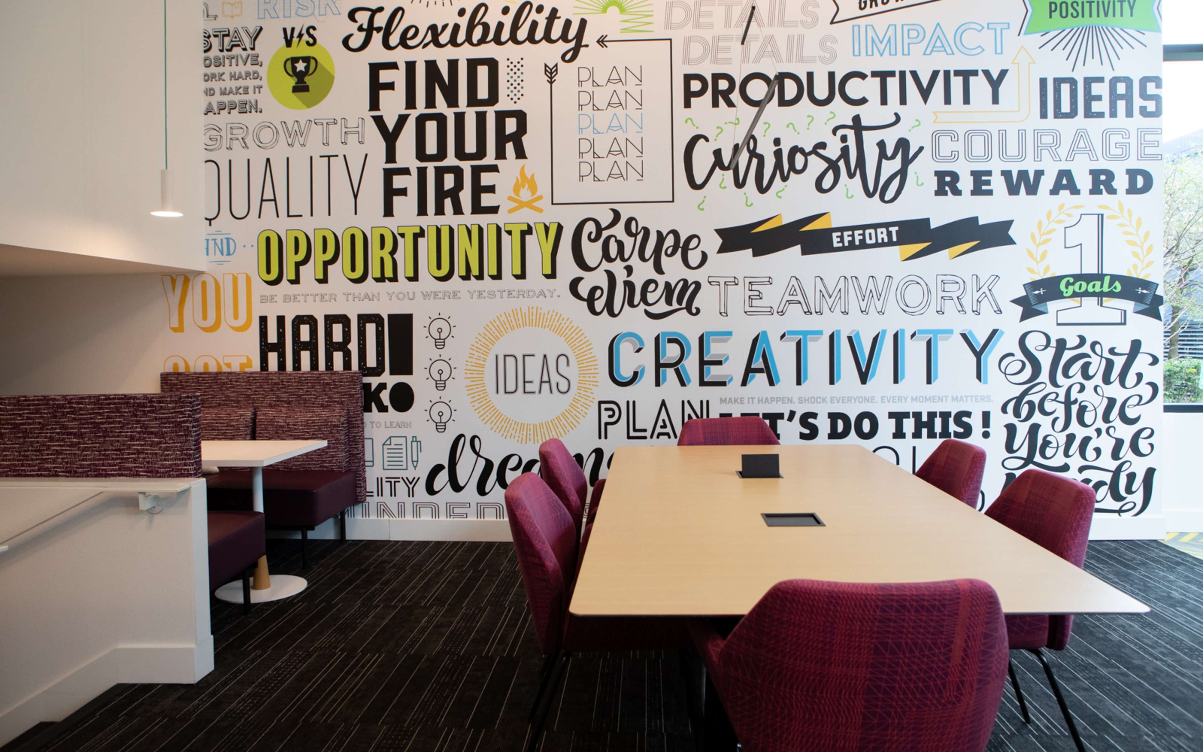 A modern meeting room with a large table surrounded by red chairs, featuring a wall covered in motivational words and phrases.