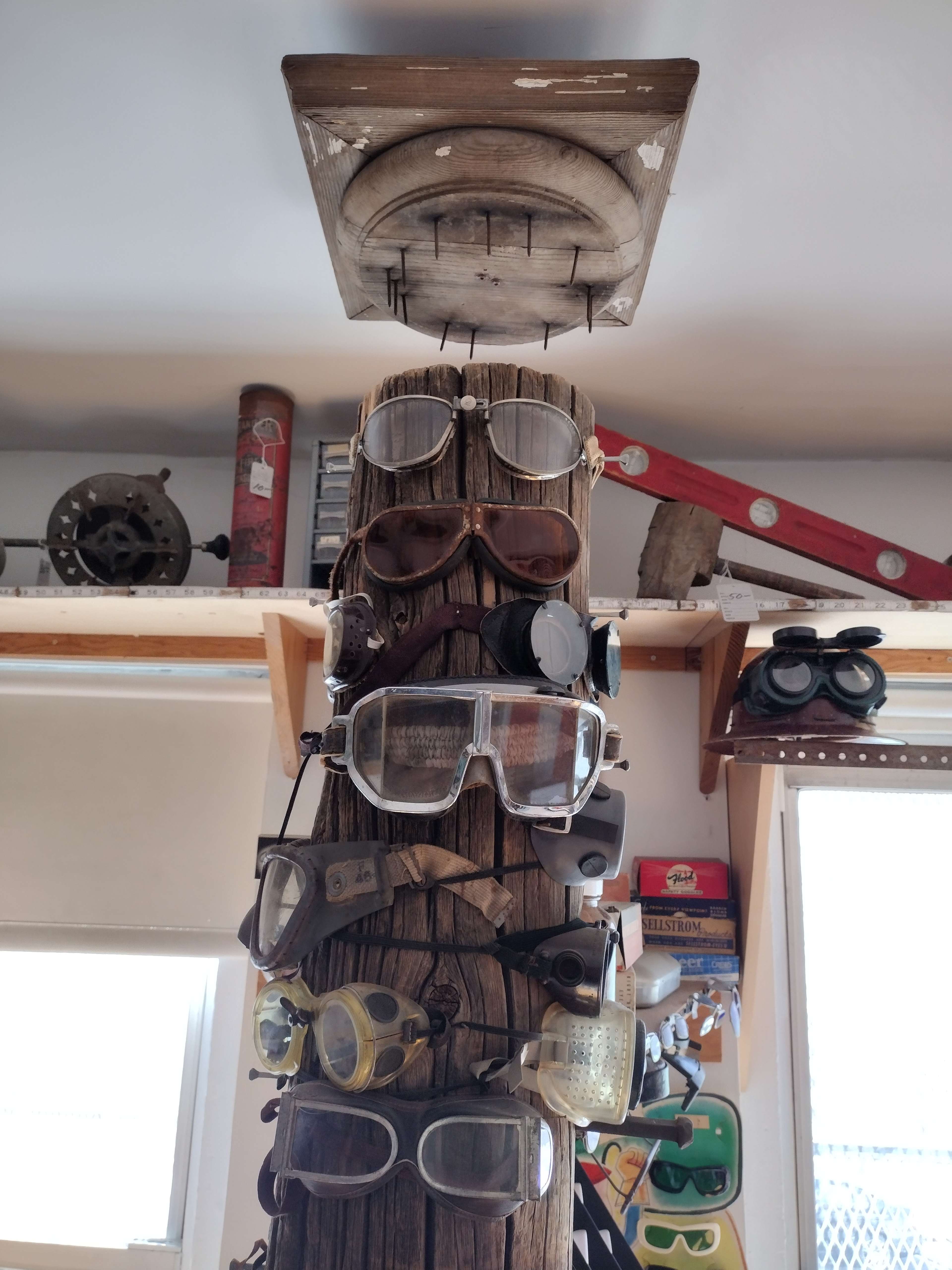 The image shows a wooden post displaying a collection of various types of goggles and glasses arranged vertically.