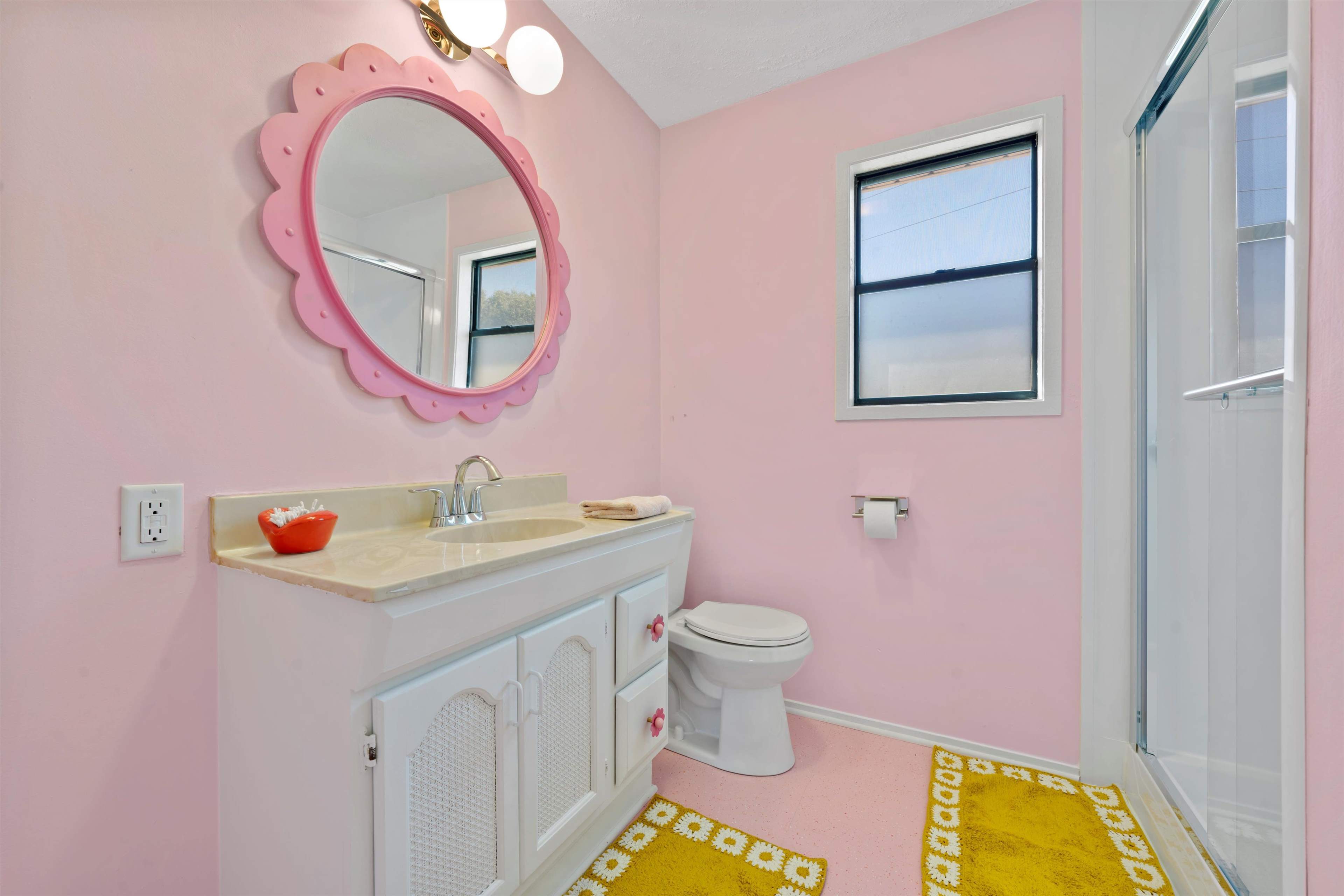 A bathroom with pink walls, a white vanity with a mirror, a toilet, and a shower area.