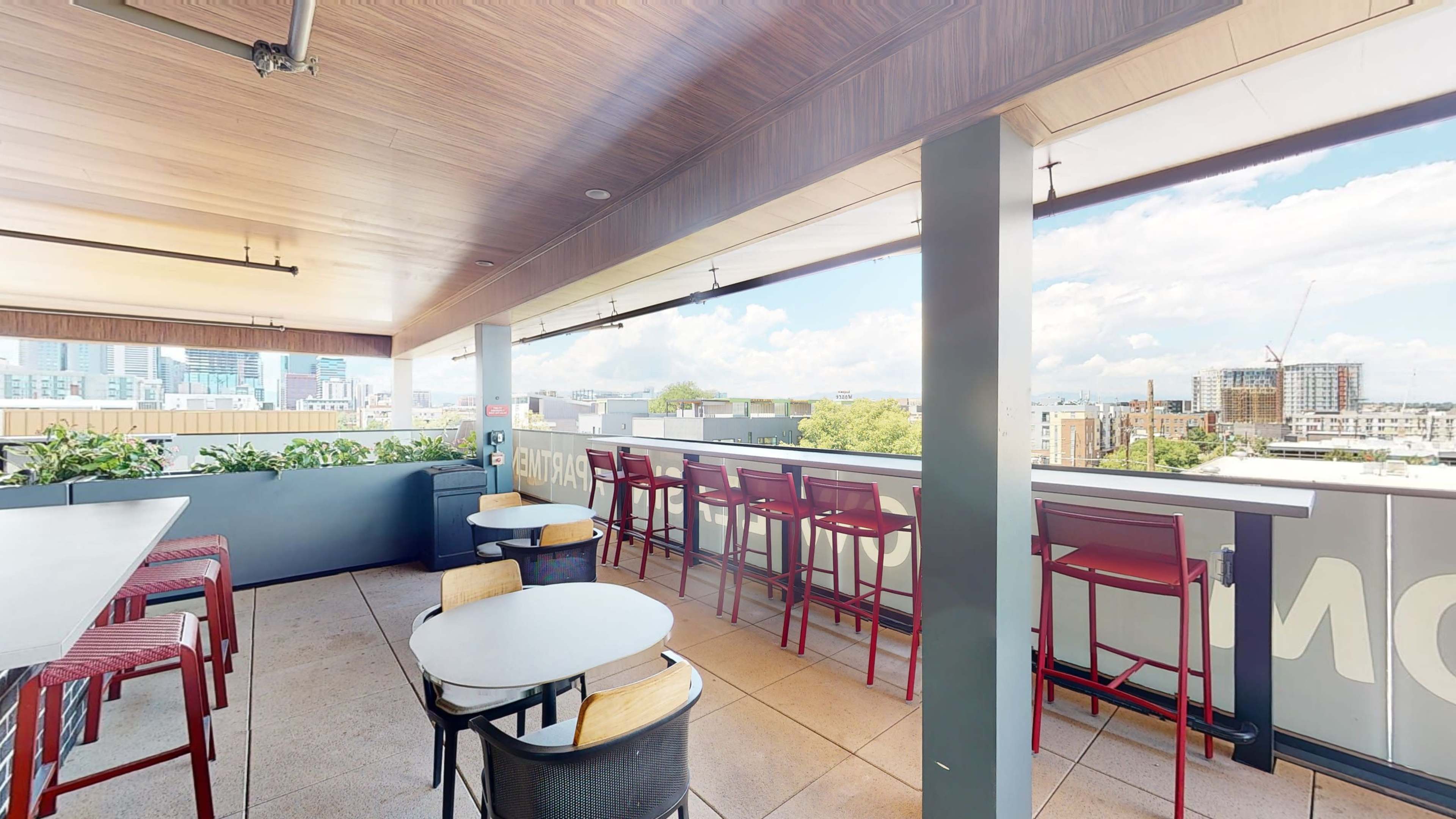 The image shows a rooftop bar with several tables and tall chairs, overlooking a cityscape with a partly cloudy sky.