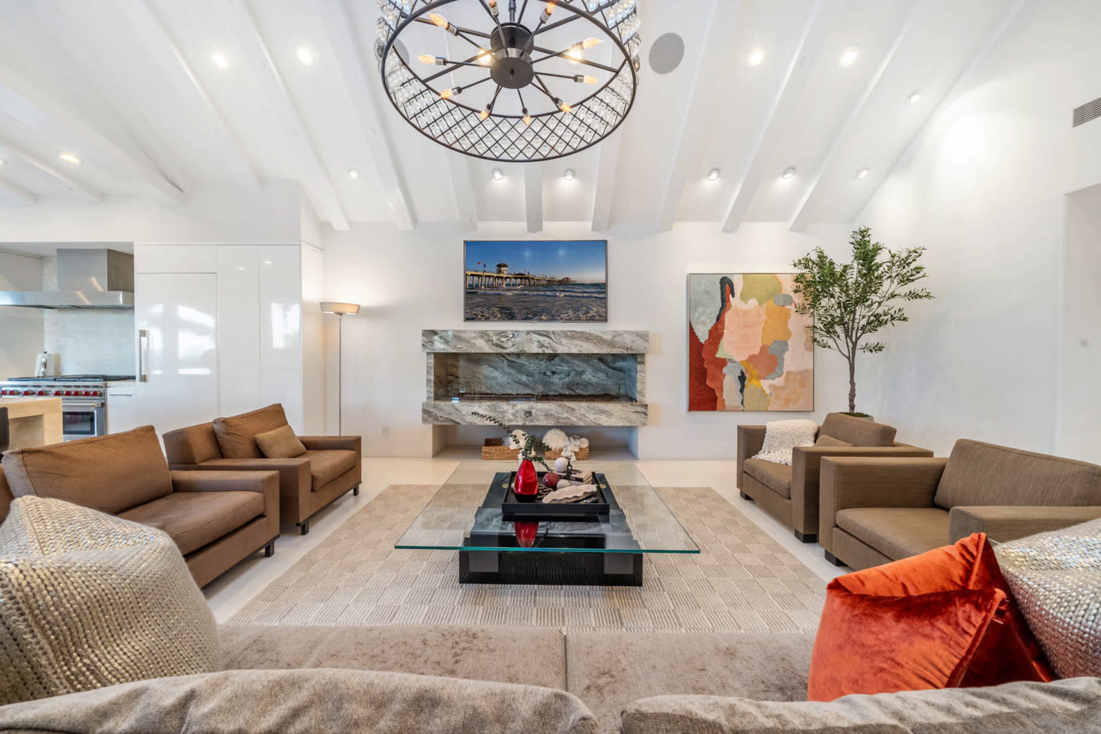 The image shows a modern living room featuring a charcoal and glass coffee table, several beige couches, a large artwork on the wall, and a decorative plant beside a fireplace.