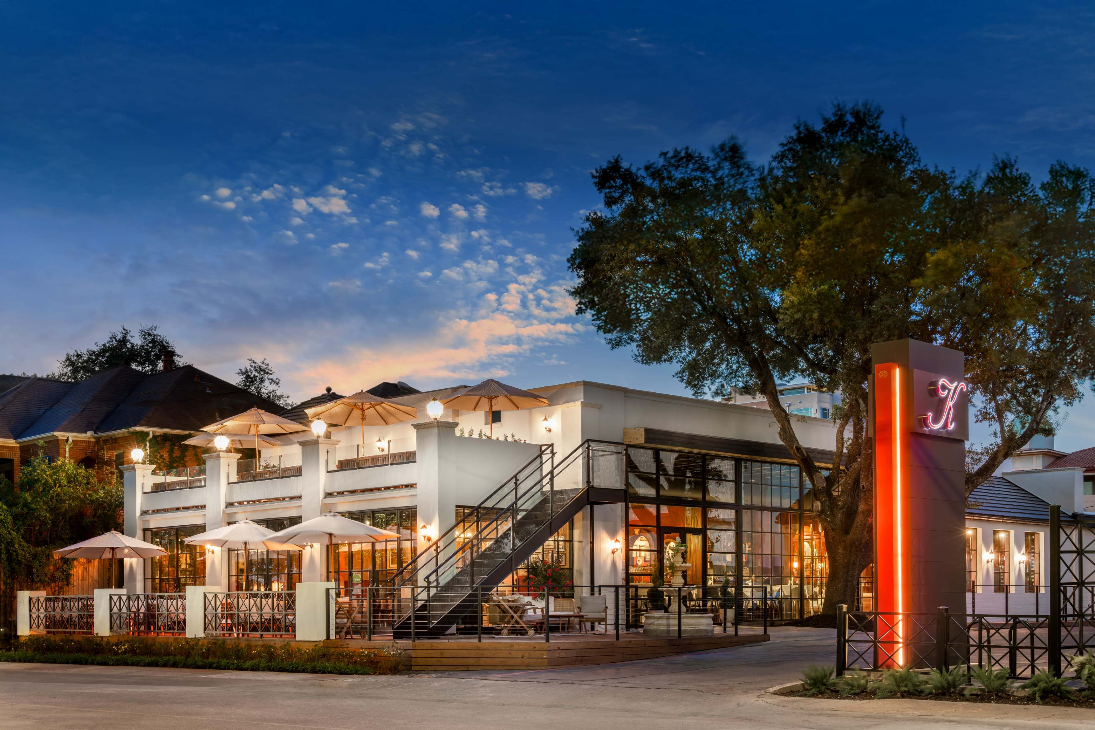 The image shows a modern two-story restaurant with large windows, outdoor seating, and a prominent sign in front, set against a twilight sky.