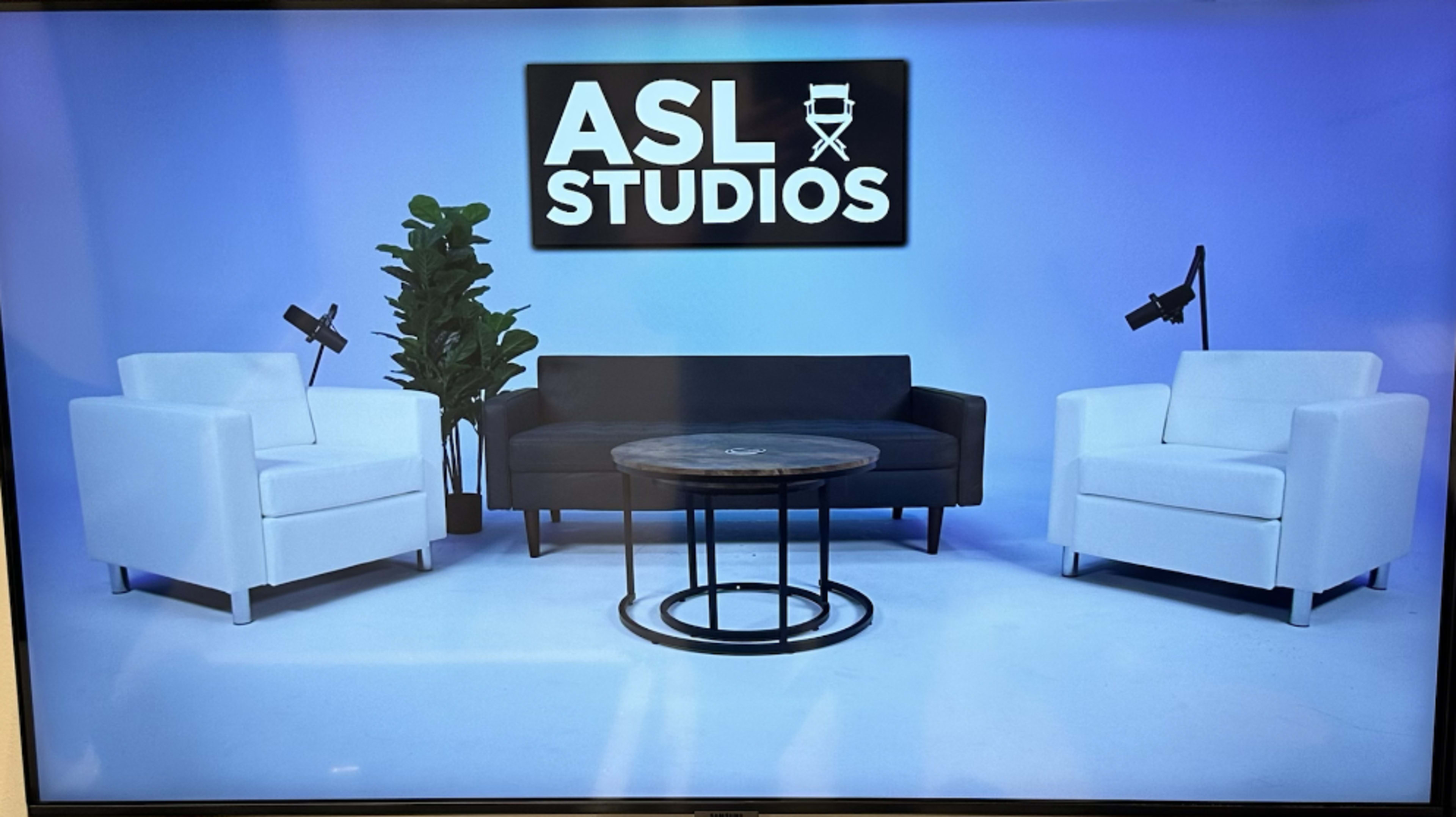 A minimalist studio setting with two white armchairs and a black sofa around a circular table, accompanied by a plant and two overhead lights.