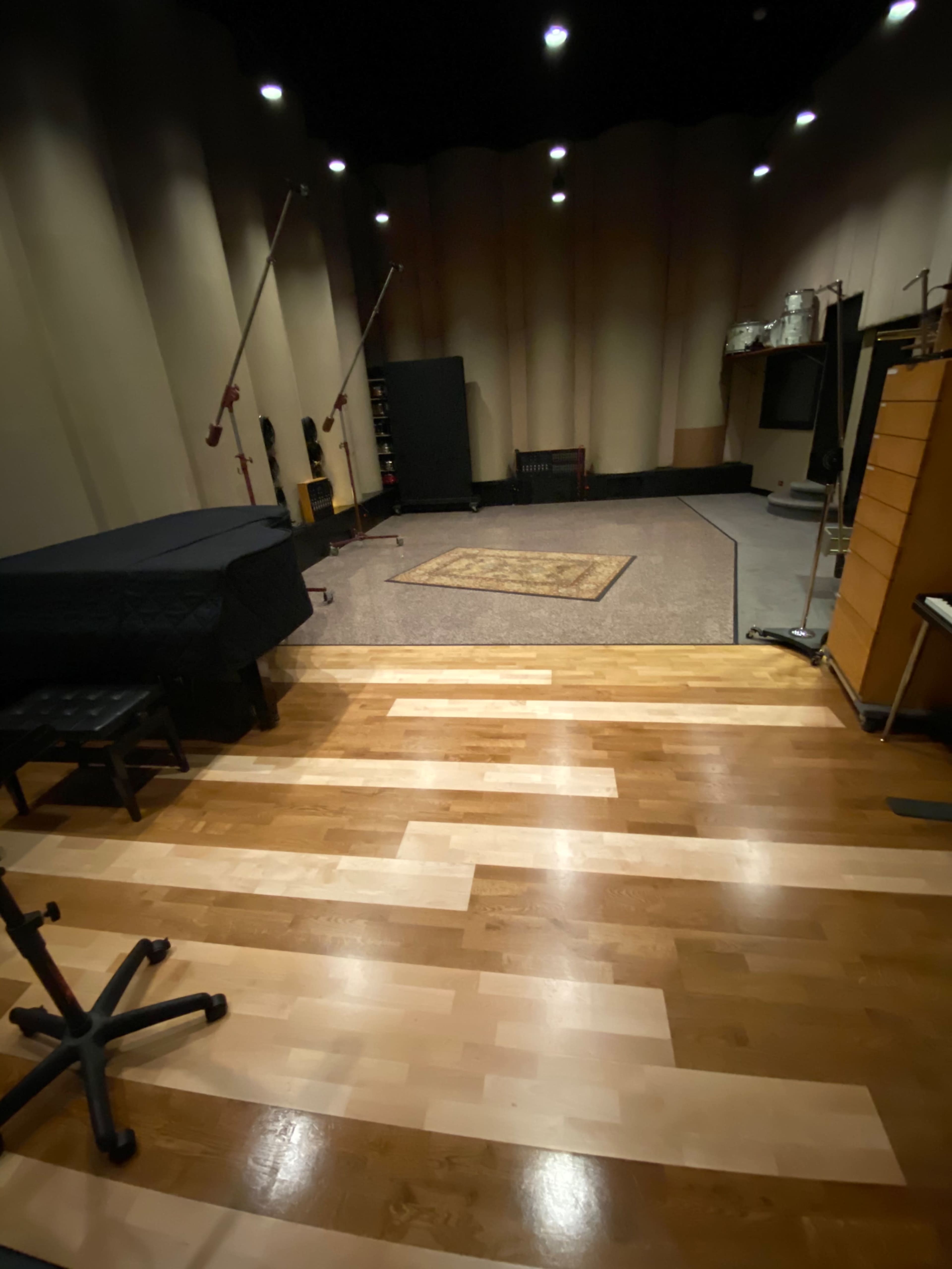 A spacious recording studio with wooden flooring, a large area rug, and various sound equipment positioned along the walls.