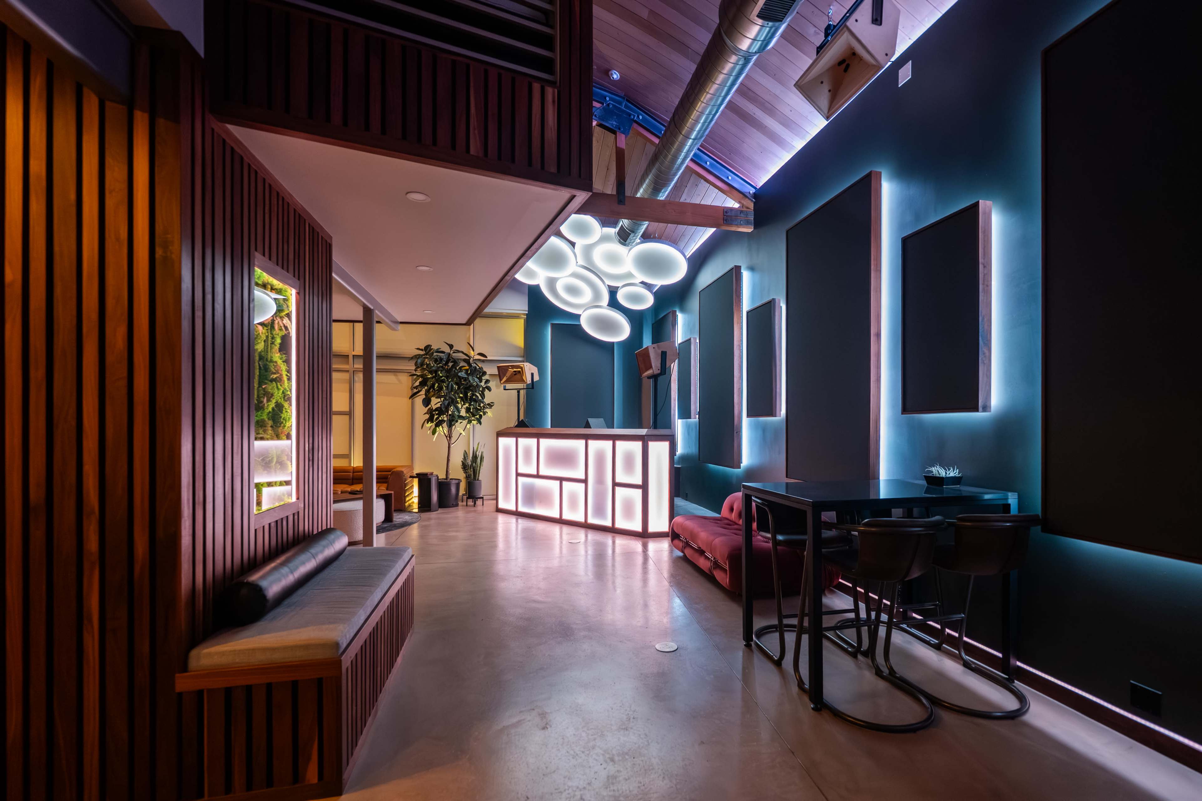 A modern interior features wooden paneling, a sleek bar with backlighting, and contemporary seating arrangements under circular light fixtures.
