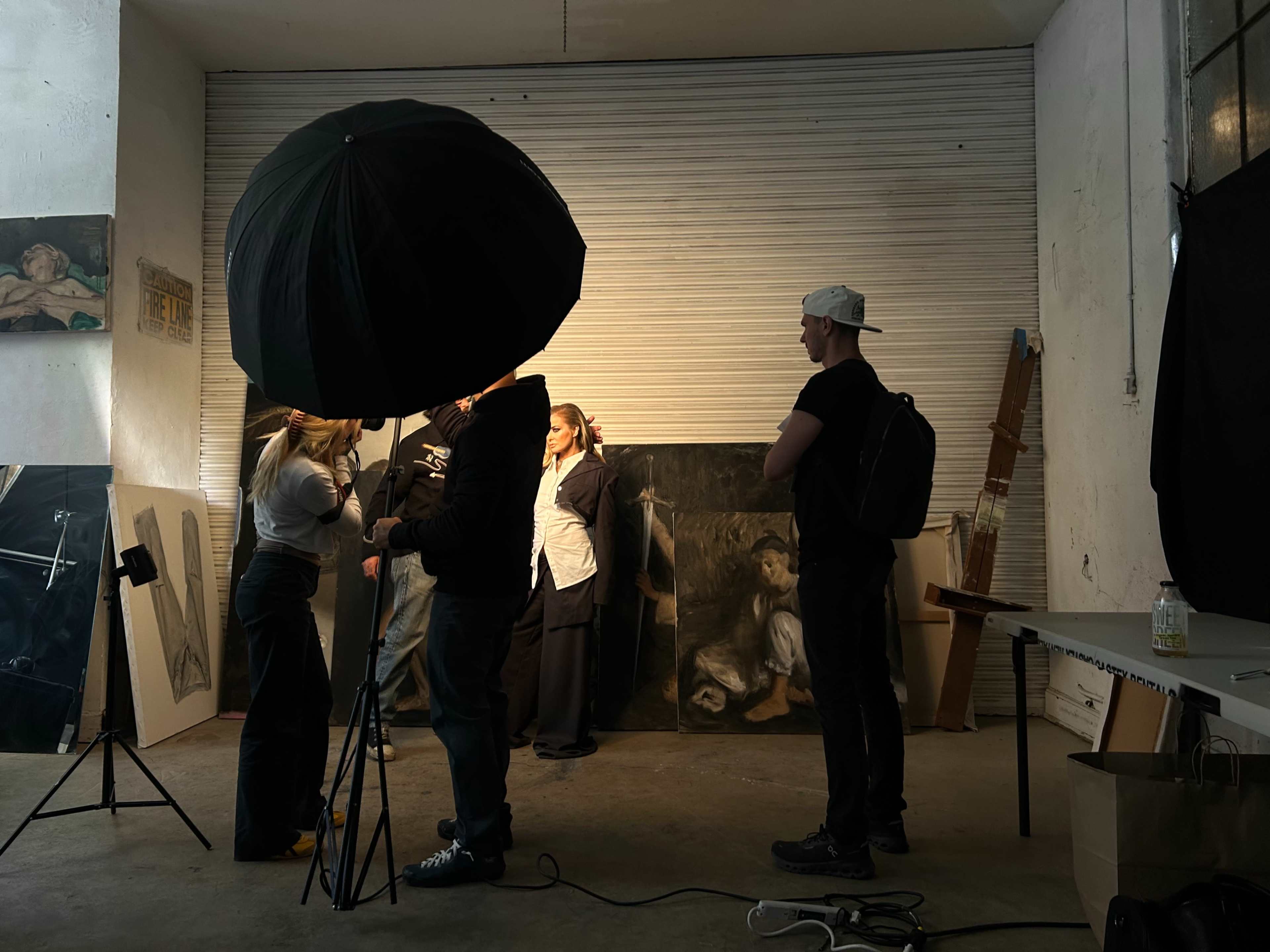 A group of people is gathered in an art studio, with one person holding an umbrella light near a backdrop of paintings.