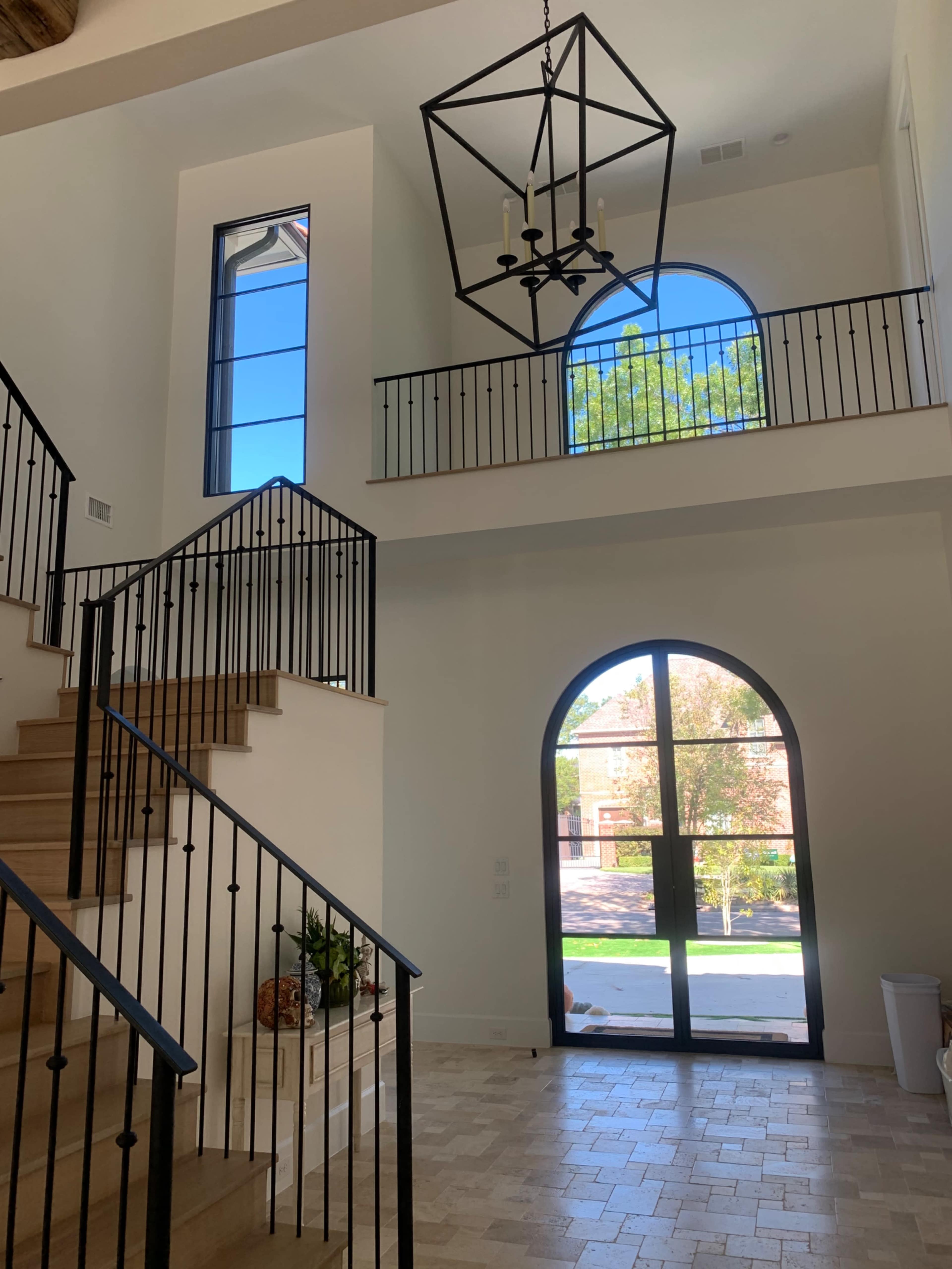 The image shows a spacious two-story foyer with a staircase, a large arched doorway, and a geometric chandelier hanging from the ceiling.