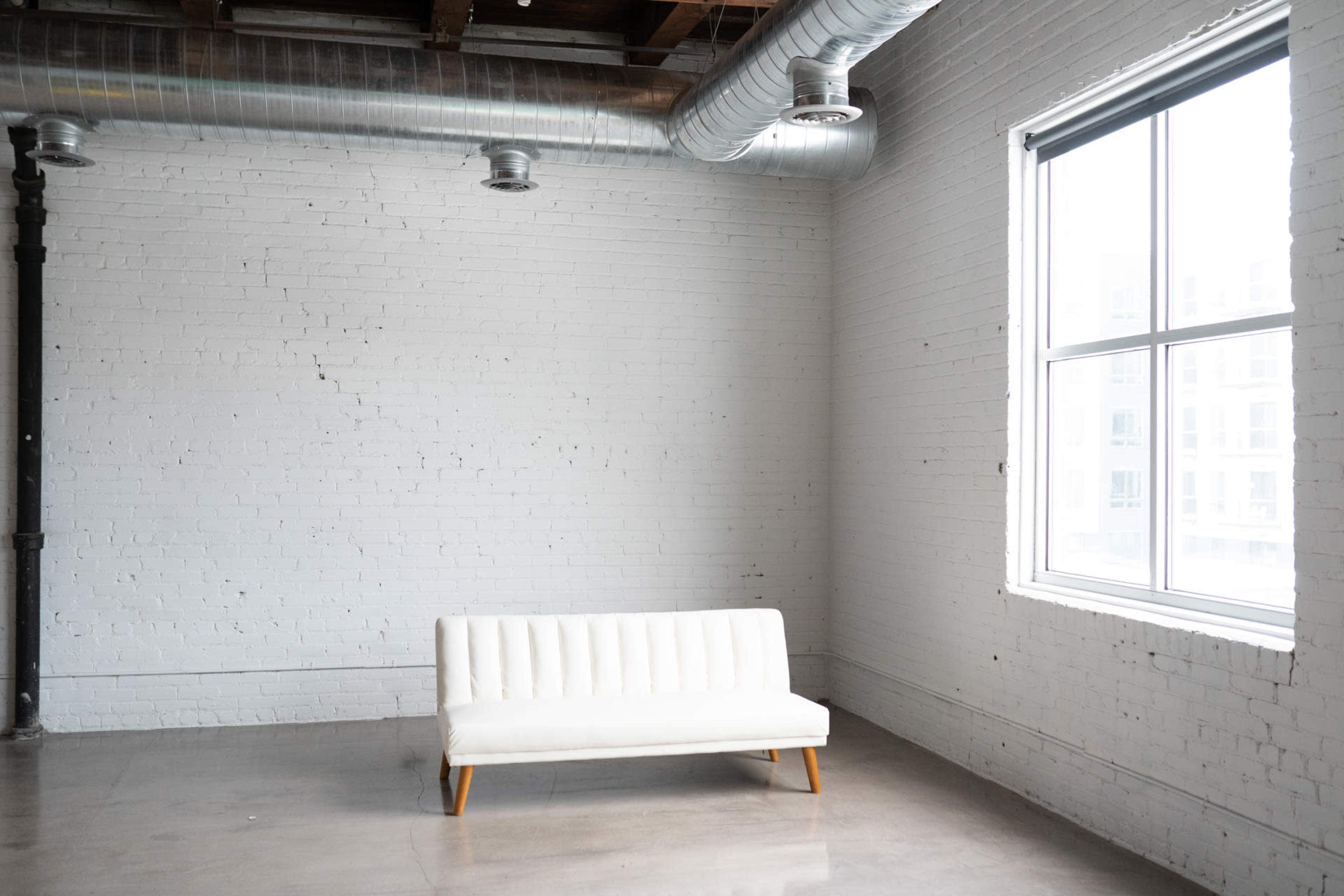 A white sofa is positioned against a light gray brick wall in a spacious, minimally decorated room with large windows and exposed ductwork.