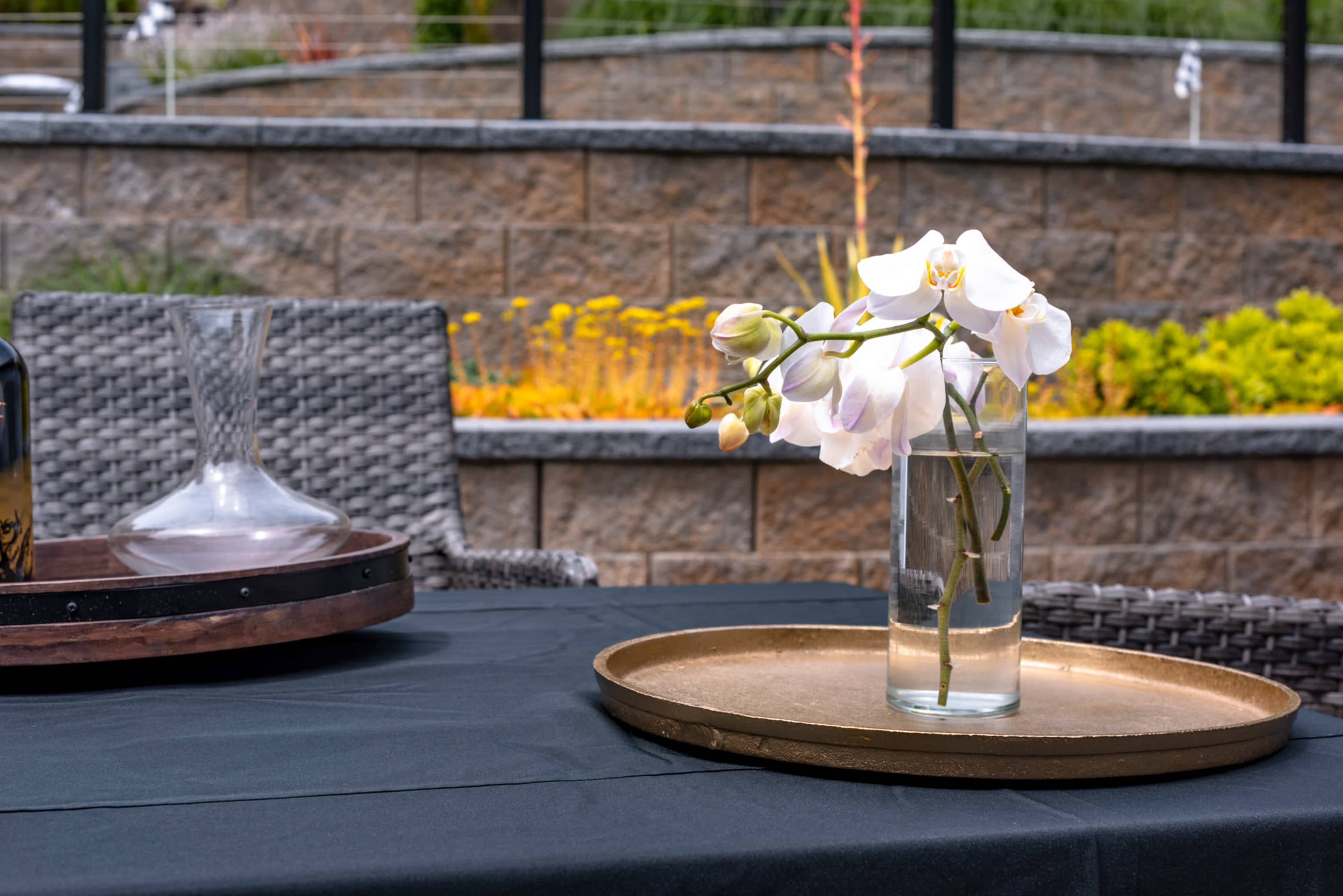 A glass vase with white orchids sits on a gold tray on a black tablecloth, with a stone wall and garden in the background.