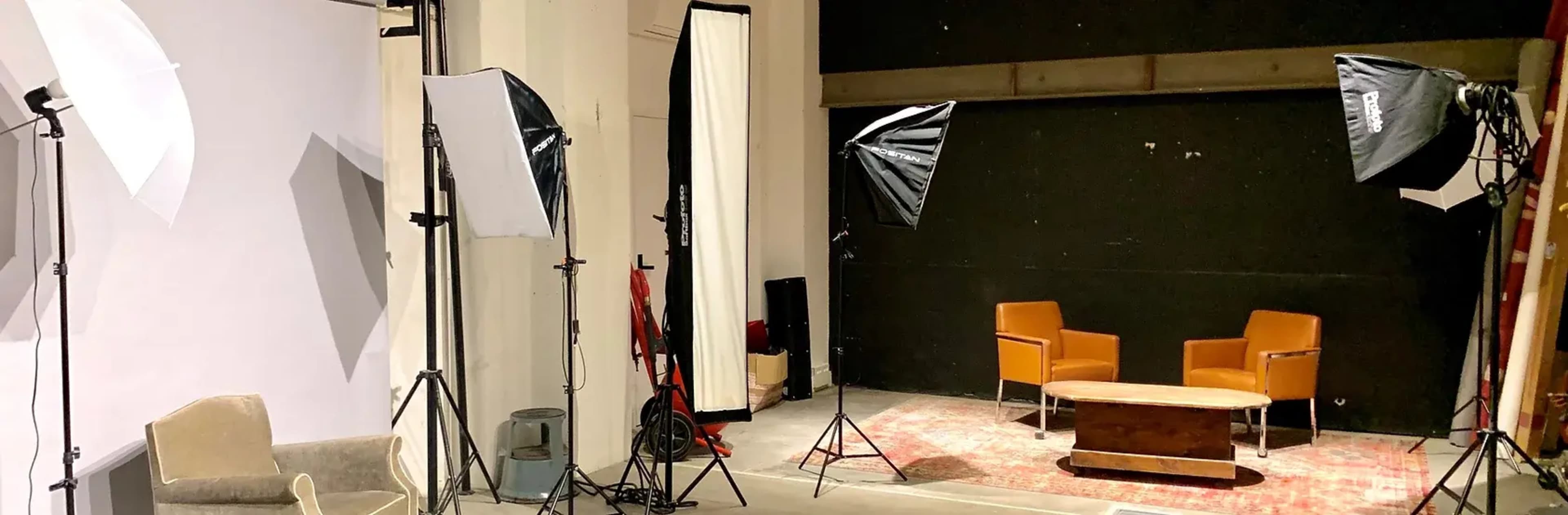 A photography studio setup features two orange chairs facing a coffee table, surrounded by lighting equipment and backdrop drapes.