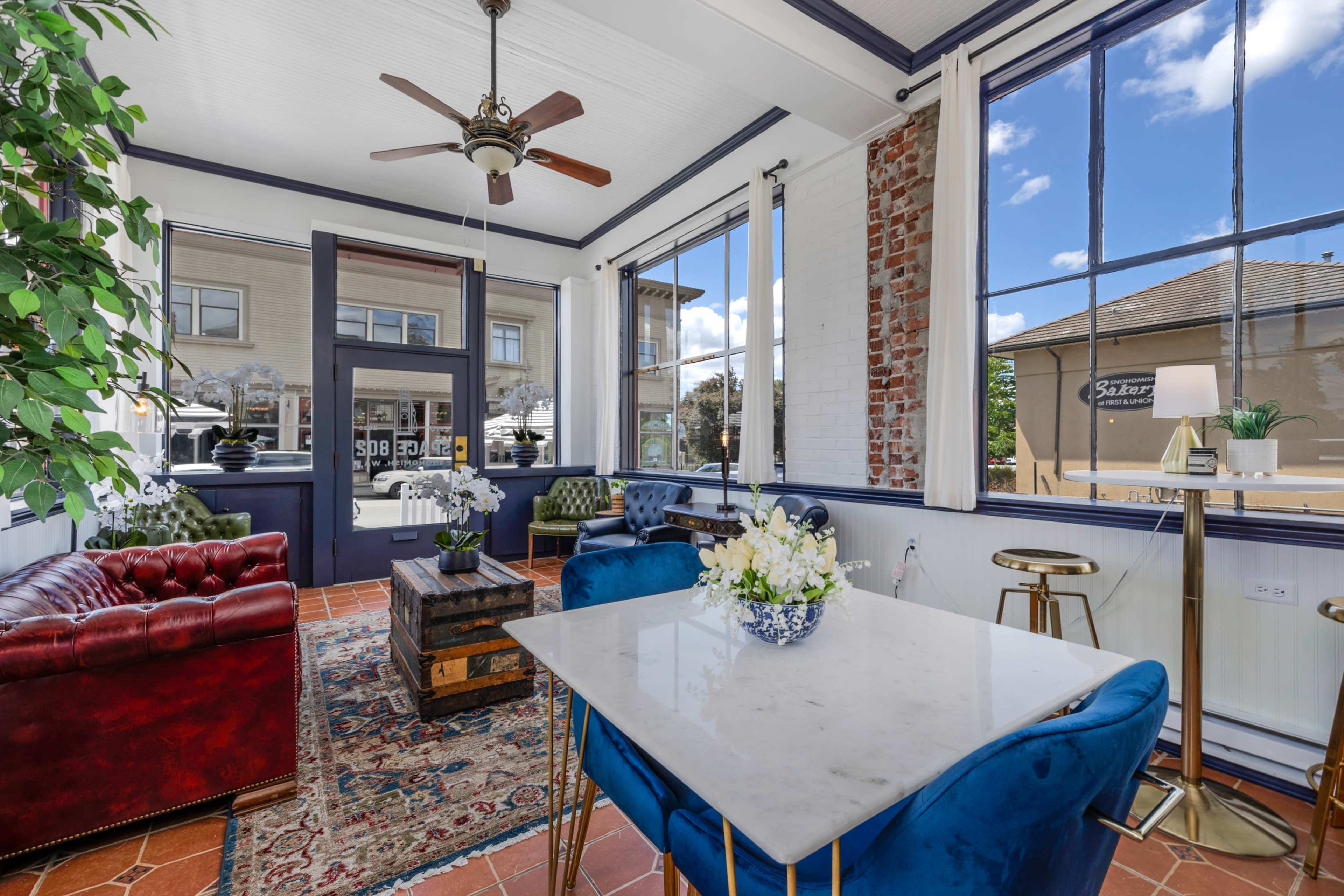 The image shows a stylish seating area with a marble table, blue armchairs, and large windows allowing natural light to enter, creating an inviting atmosphere.