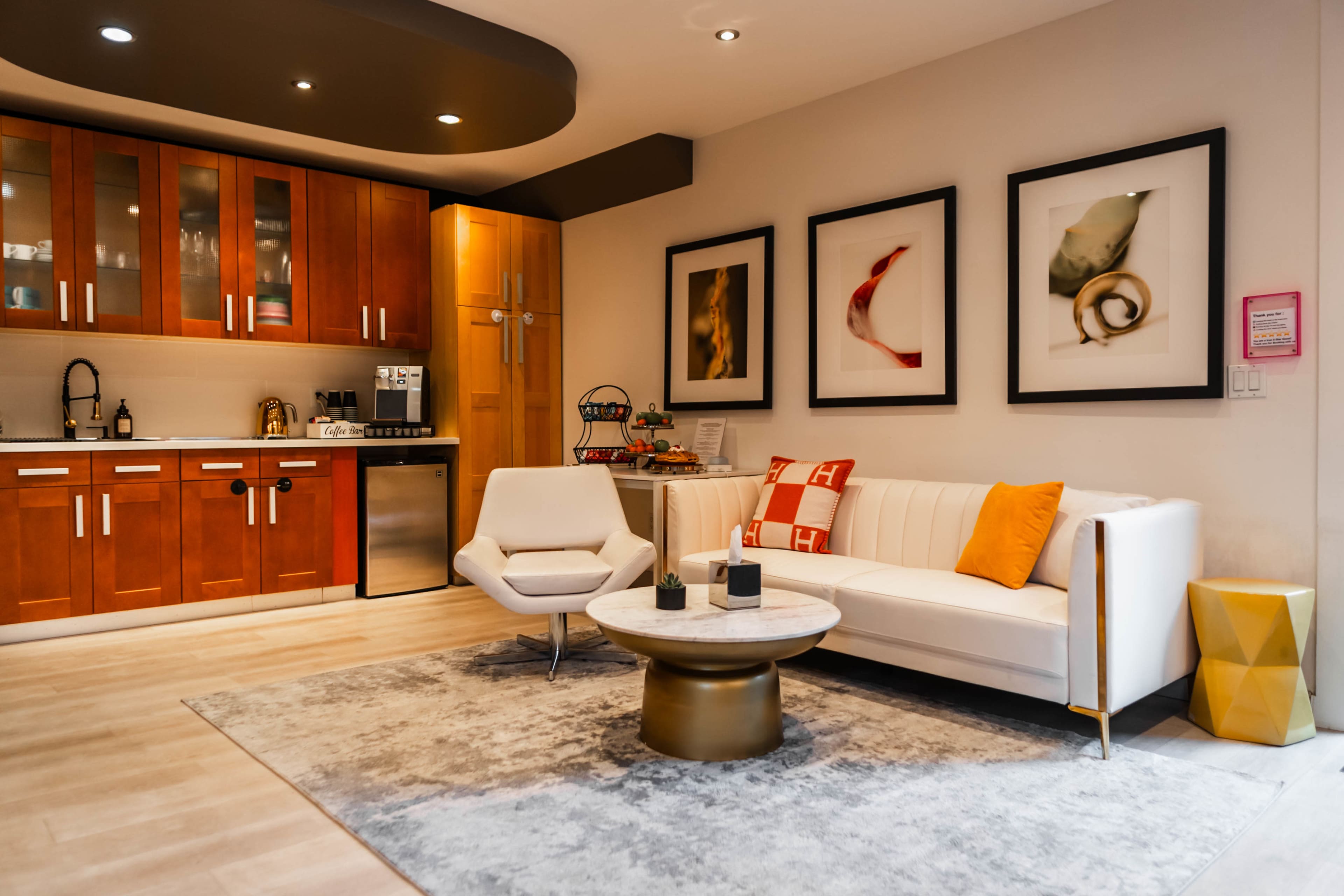 A modern living space featuring a kitchen area with wooden cabinets, a white sofa with colorful cushions, a round coffee table, and framed artwork on the walls.