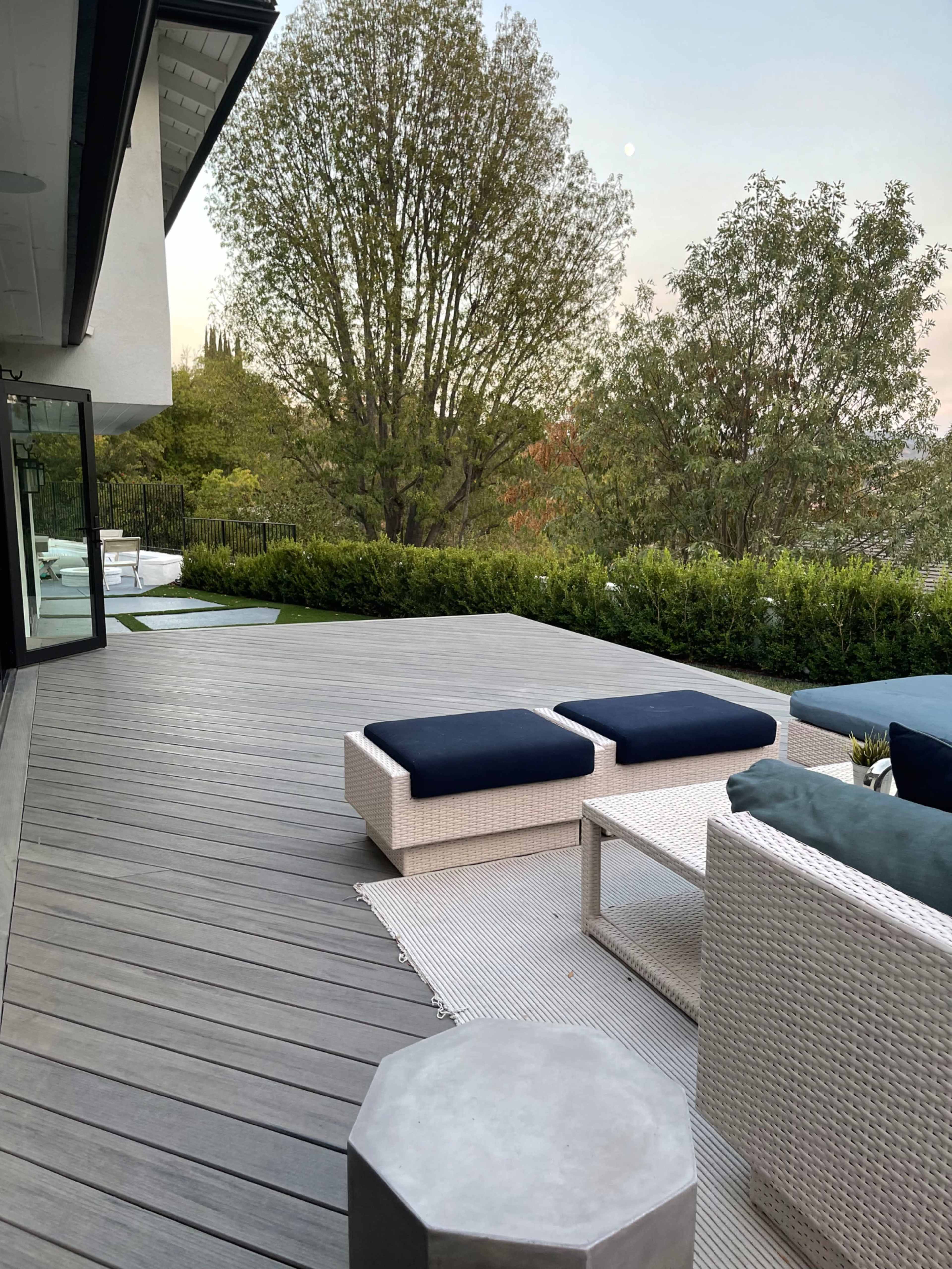 A modern patio features wooden decking, outdoor furniture including cushioned seating and a coffee table, surrounded by greenery and trees.