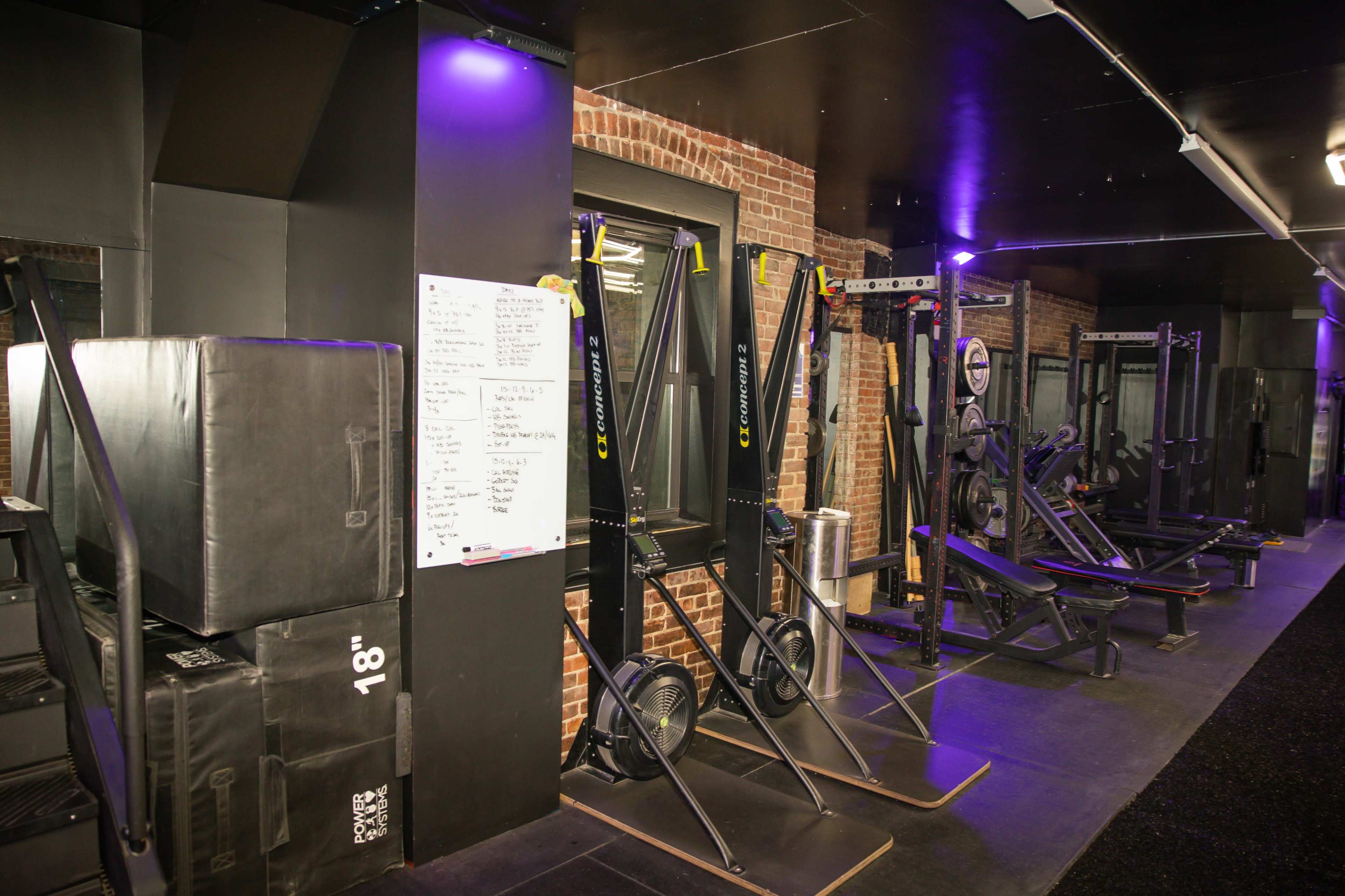 The image shows a fitness gym featuring various exercise equipment, including rowing machines, benches, and a wall of workout instructions.