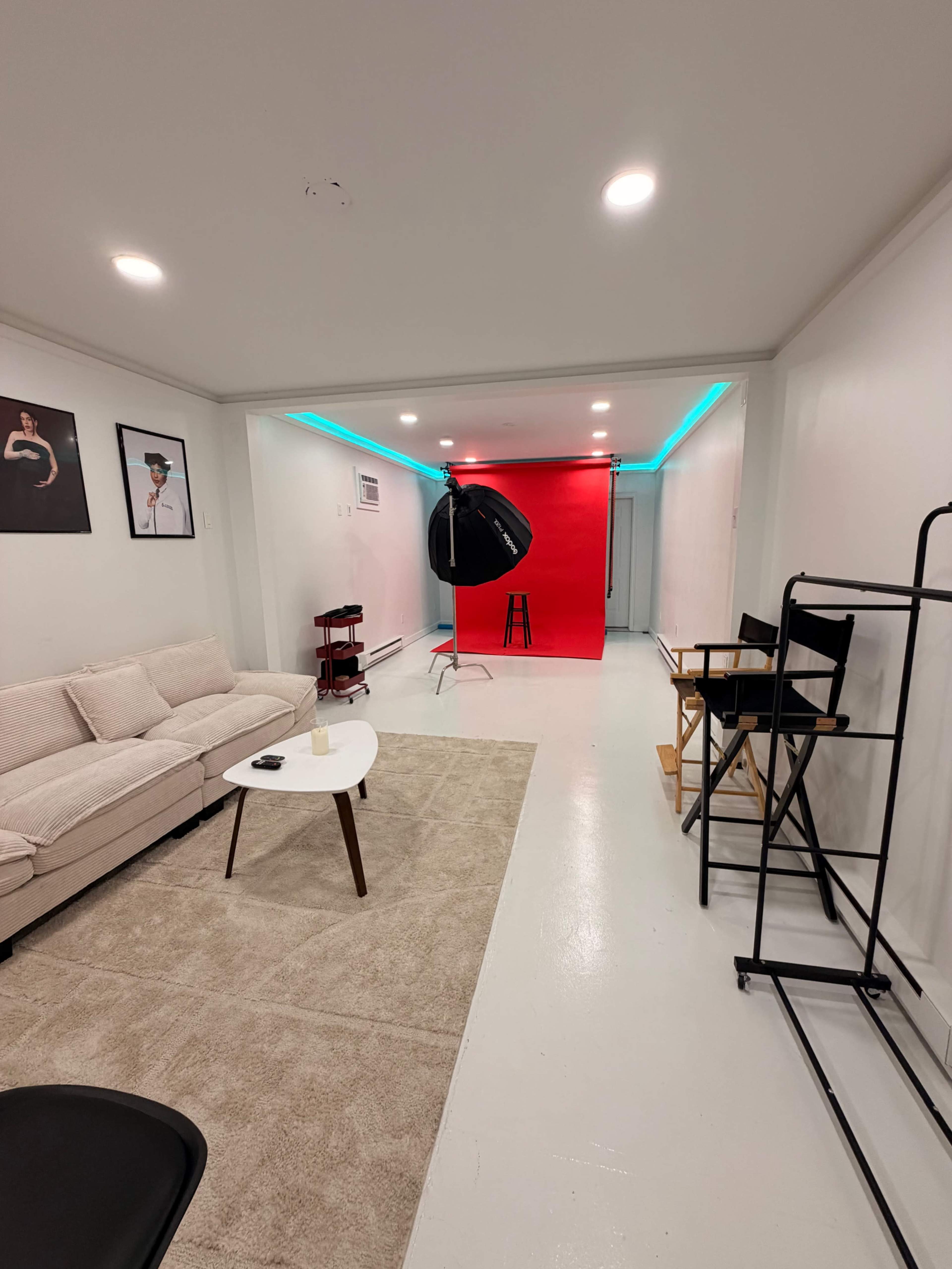 The image shows a spacious room with a beige couch, a white table, a light-colored carpet, and a backdrop of vibrant red, along with studio lighting equipment and director's chairs.
