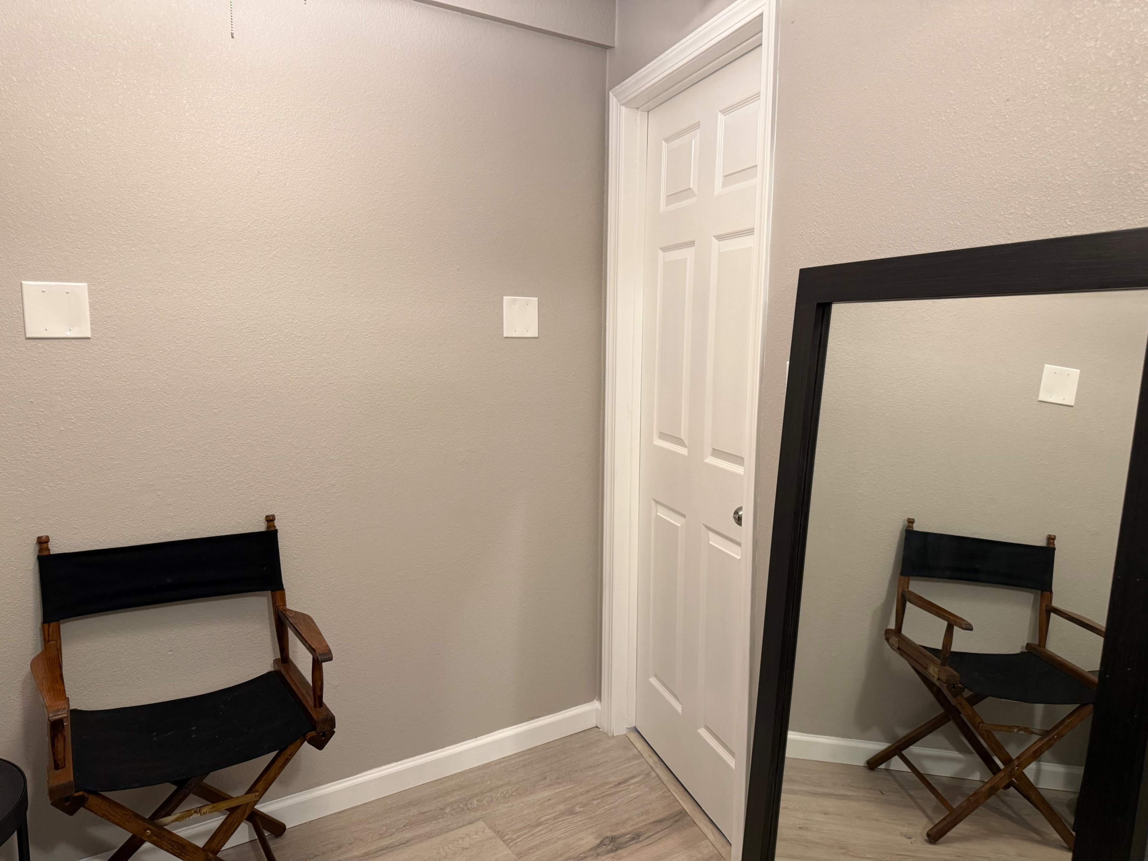 A small room features a wooden folding chair beside a mirrored wall and a closed white door.
