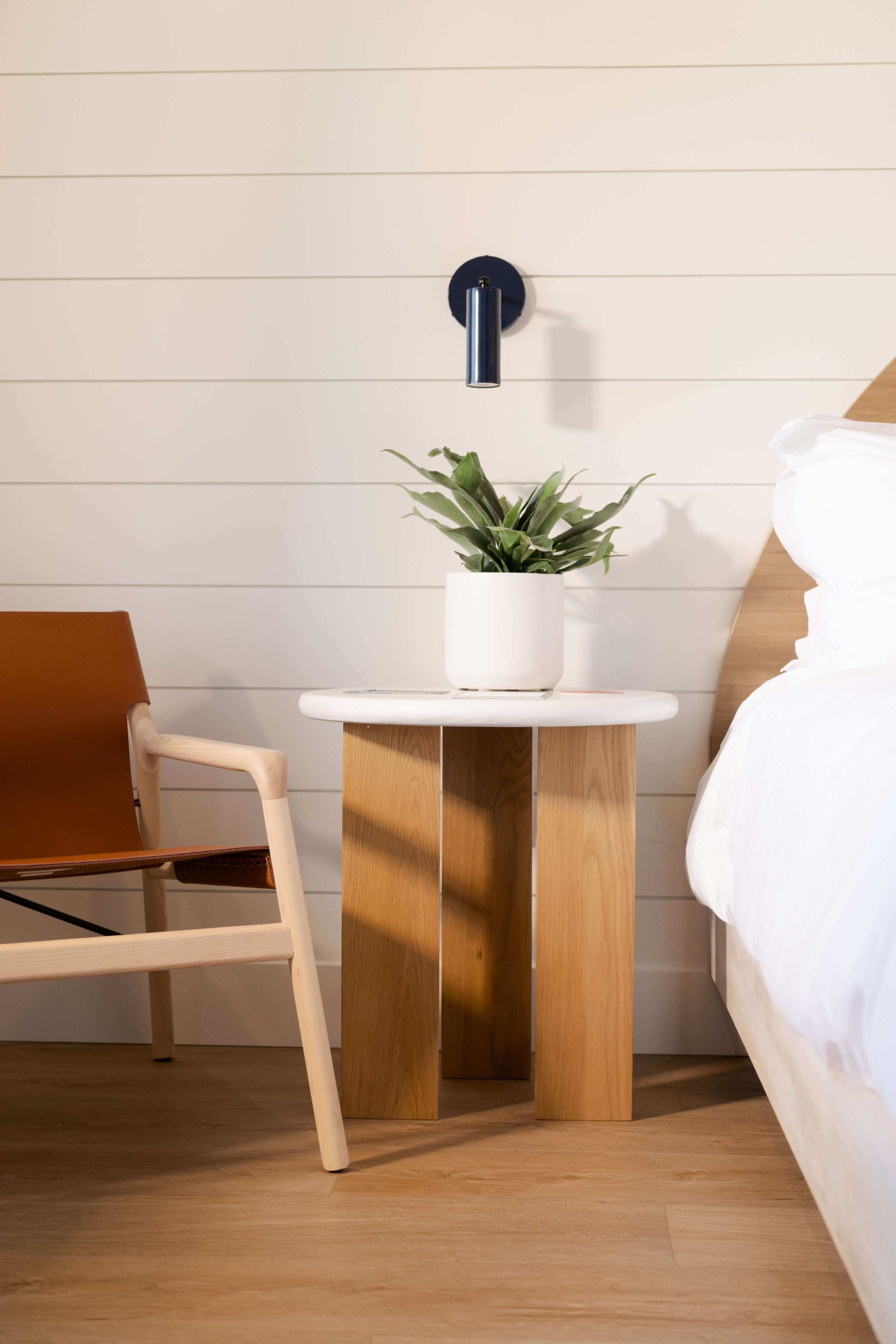 A wooden side table with a round marble top and a potted plant is positioned next to a bed with white linens and a brown armchair nearby.