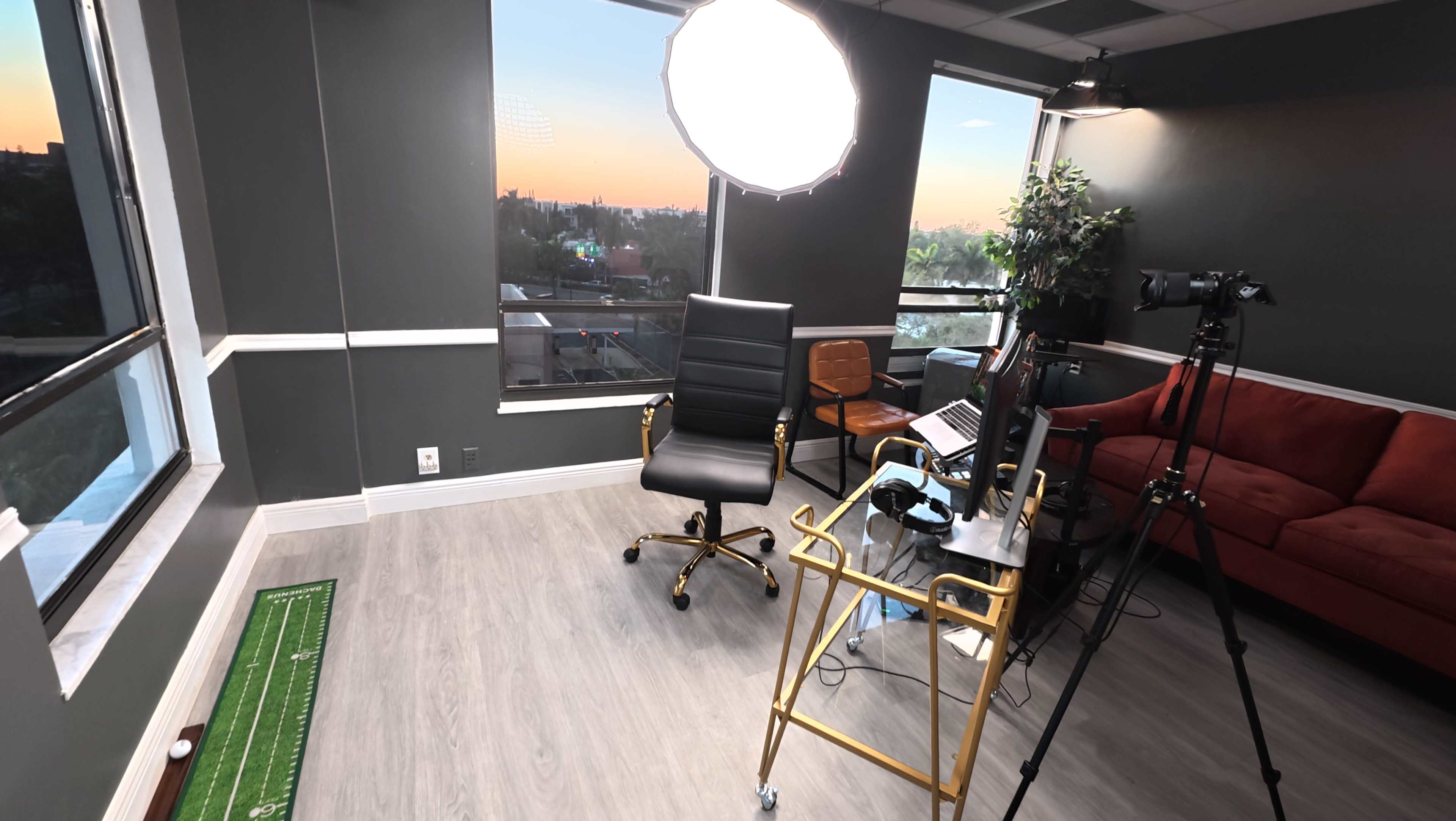 The image shows a modern office space with a large window, a black swivel chair, a brown armchair, a small table, and a lighting setup for filming or photography.