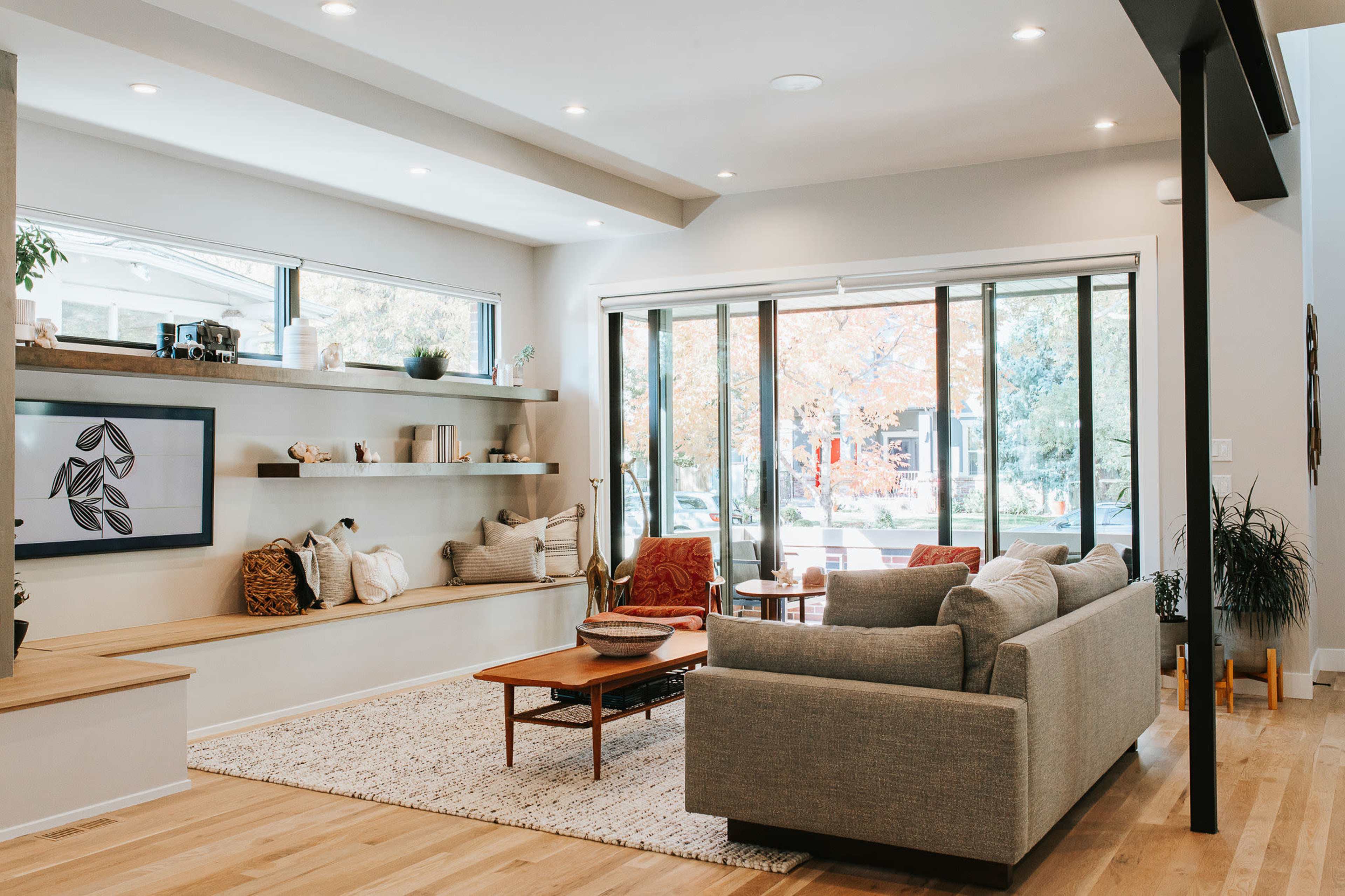 The image shows a modern living room with large windows, a cozy seating area, and built-in shelves displaying decorative items.
