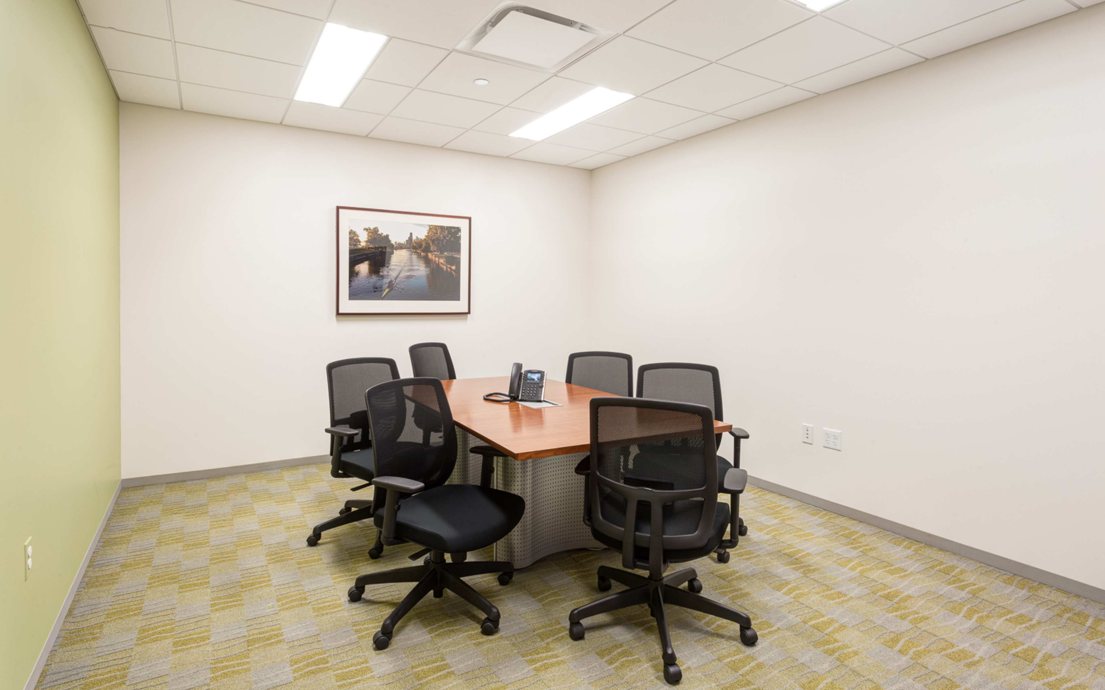 An empty conference room with a rectangular wooden table surrounded by six black mesh chairs, and a framed picture of a landscape on the wall.