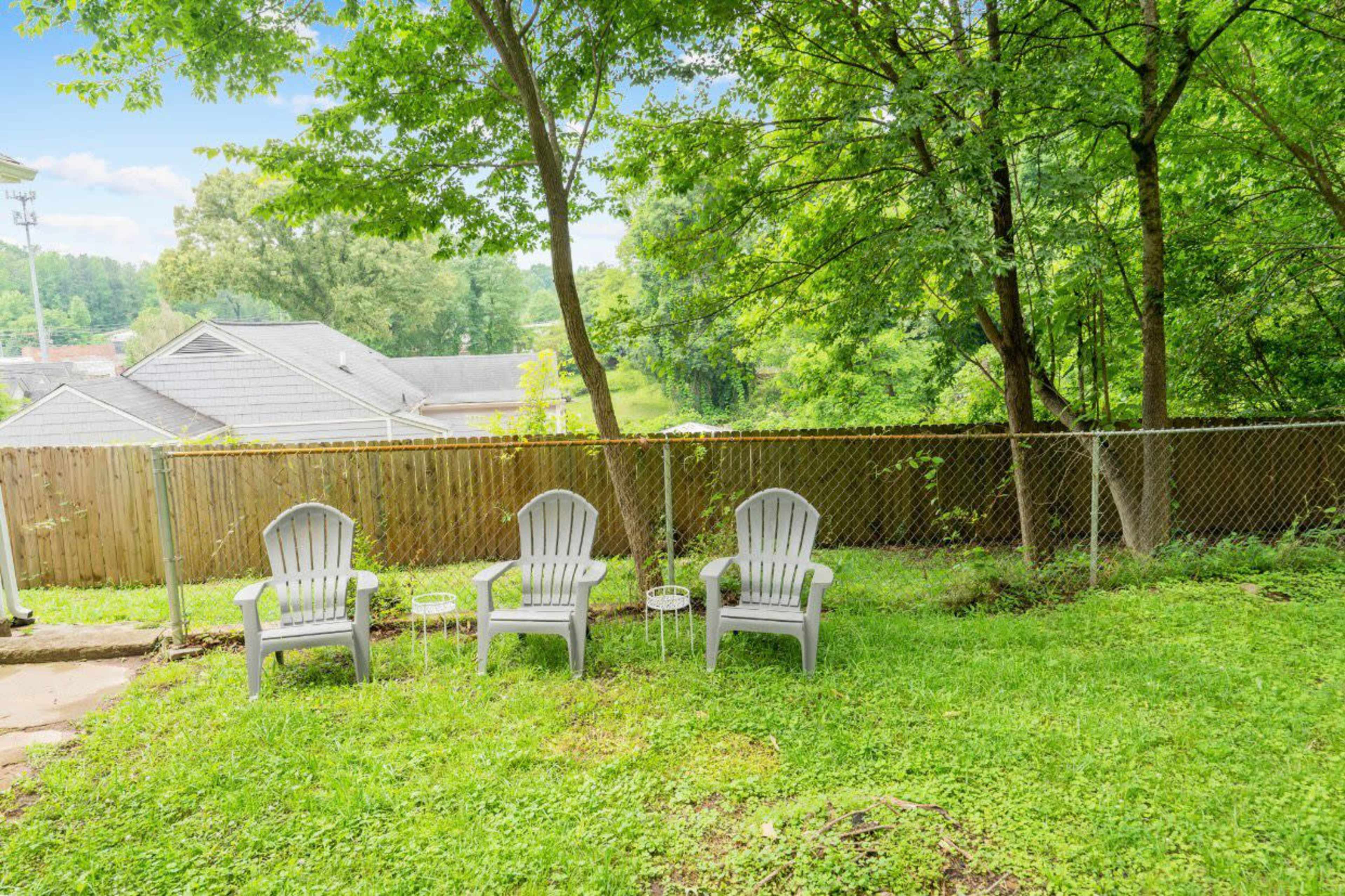 Three white Adirondack chairs arranged on a grassy area beside a chain-link fence, with trees and houses in the background.