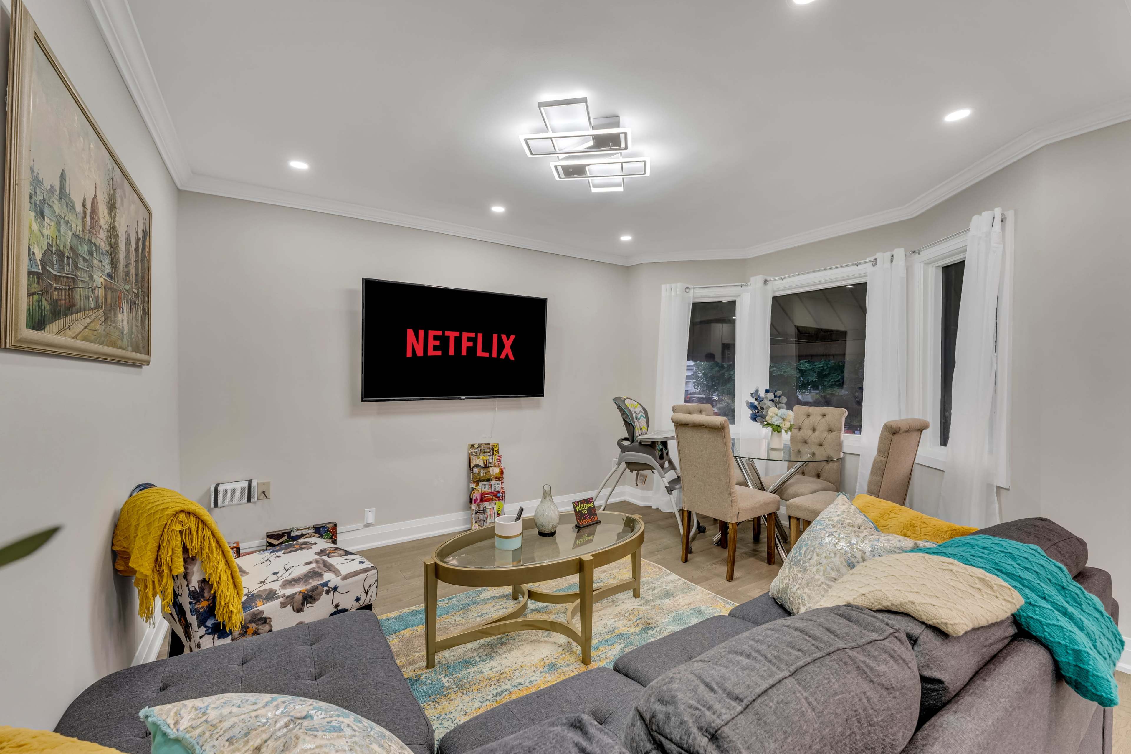 A cozy living room features a grey sofa with colorful throw pillows, a gold coffee table, and a wall-mounted TV displaying the Netflix logo, with a dining area visible in the background.