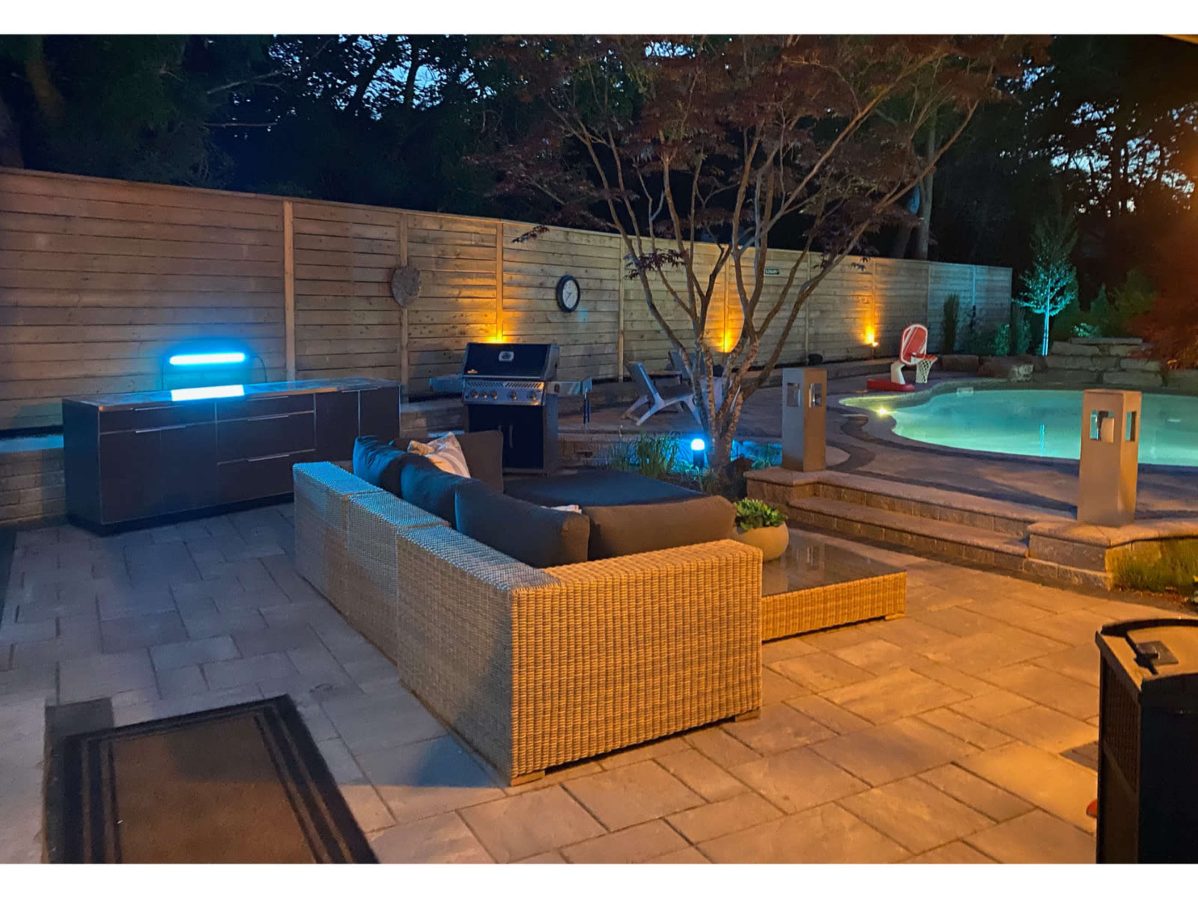 A patio area with a sectional sofa, a grill, a pool, and ambient lighting in a landscaped backyard.