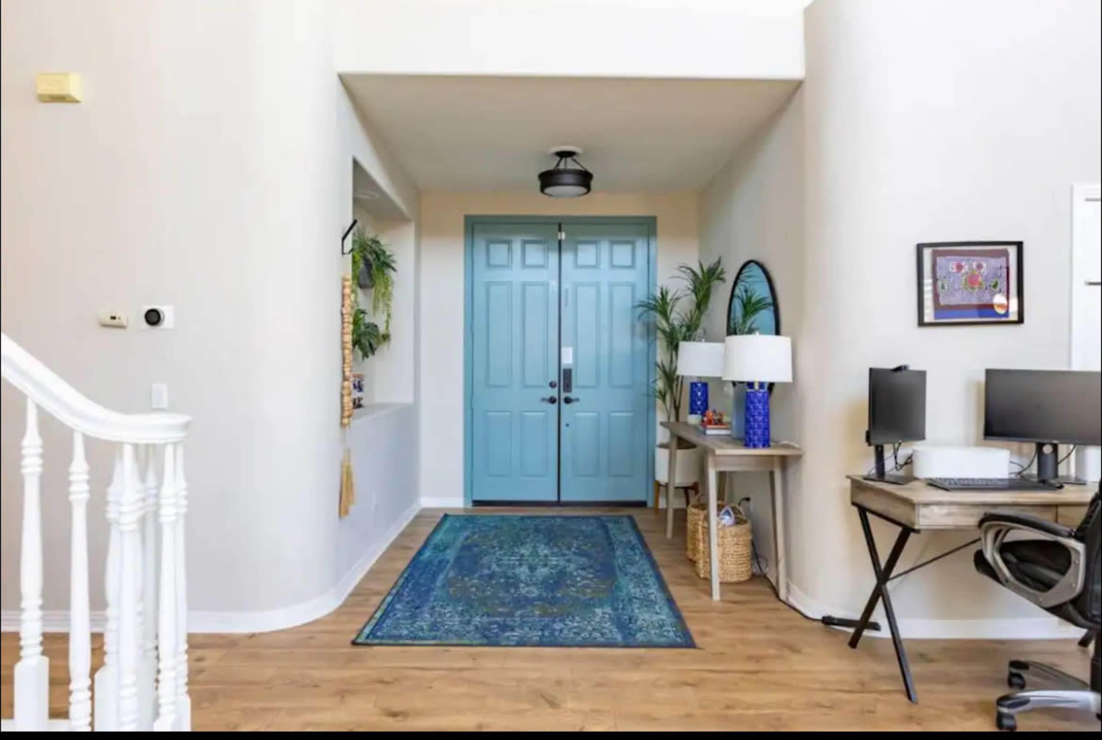 The image shows a front entryway with light-colored walls, a blue front door, a patterned rug, and a small desk with computers on one side.