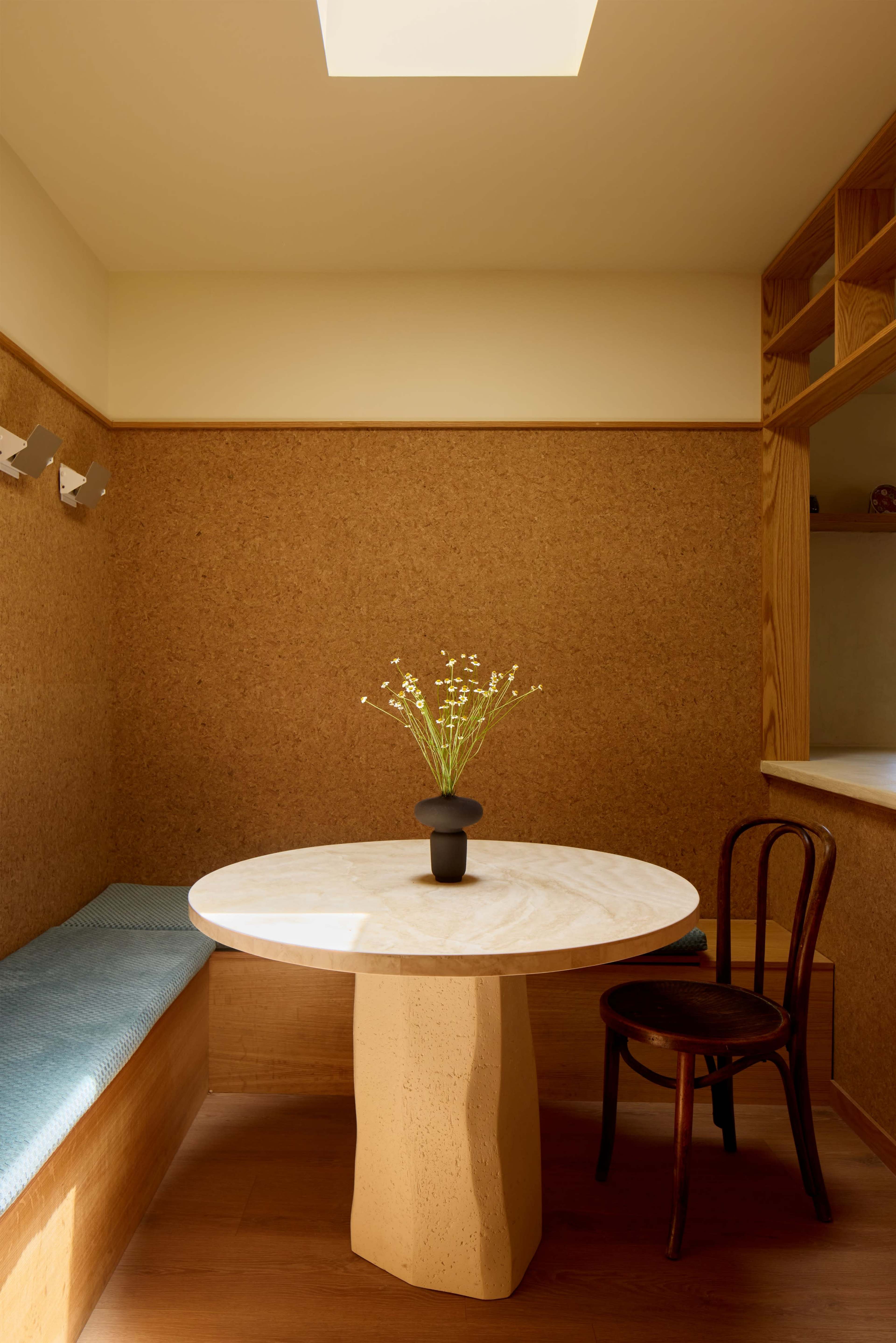 A round marble table with a stone base is set in a small room featuring cork walls and a wooden shelf, with a single black vase containing small flowers on the table.