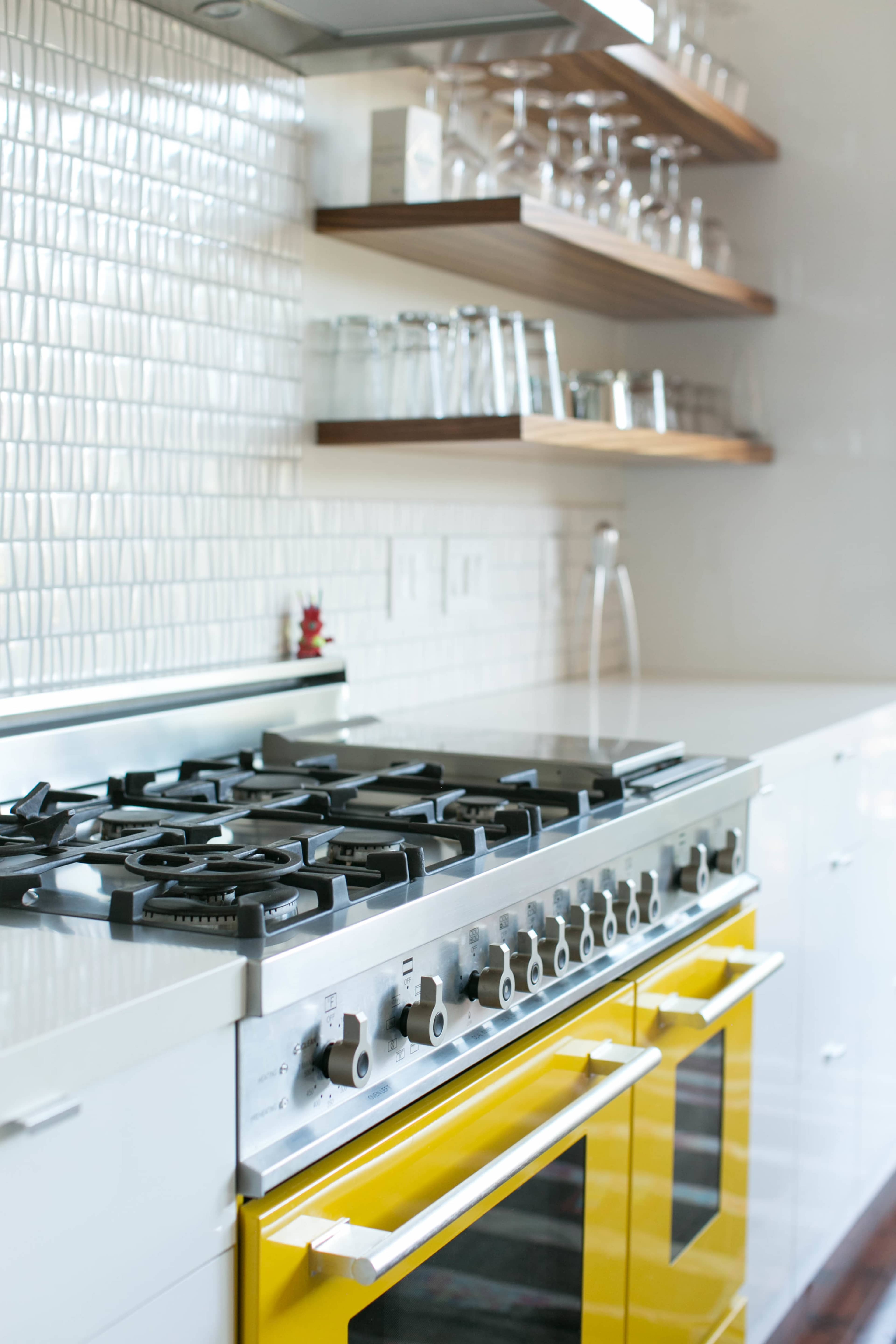 A modern kitchen with a yellow gas range and white cabinetry, featuring open wooden shelves and glassware above a tiled backsplash.