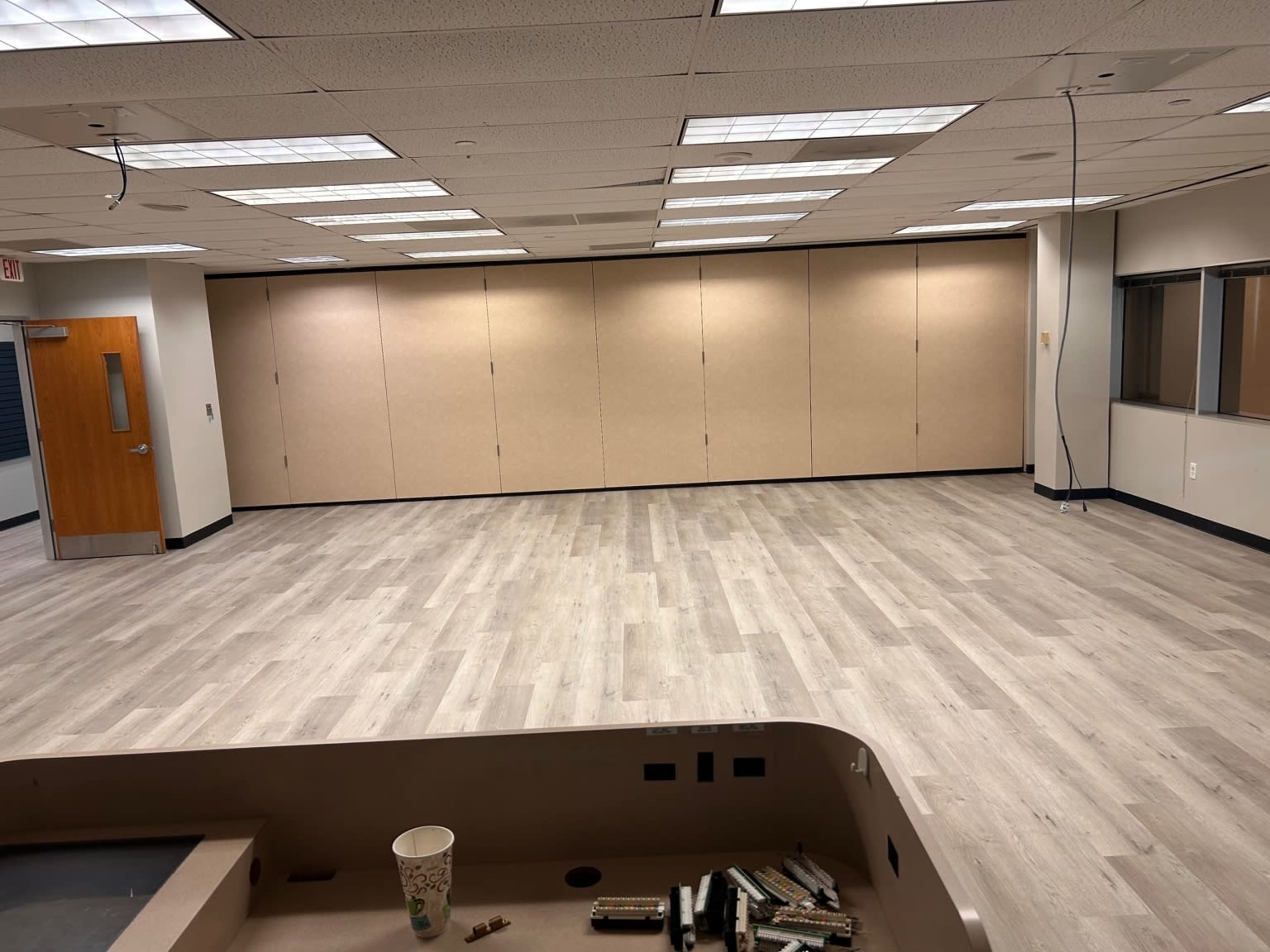 The image shows a nearly empty office space with light wood flooring, beige wall panels, and overhead lighting.