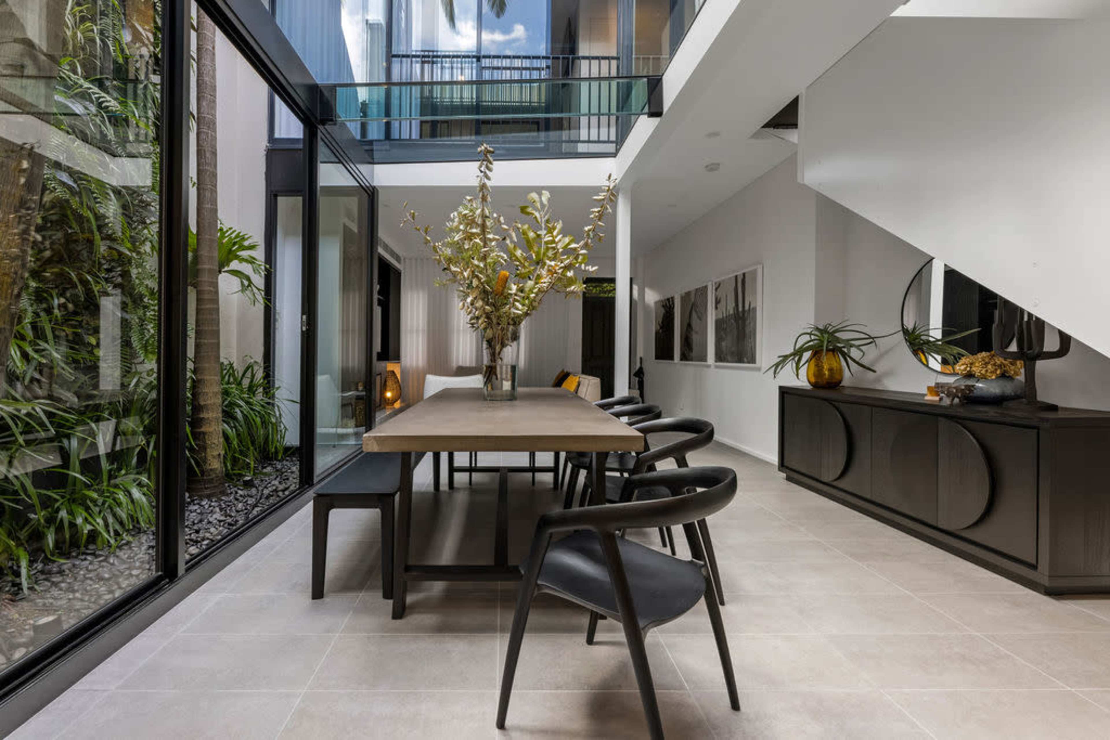 A modern dining area with a long wooden table, black chairs, and large windows that overlook a green outdoor space.